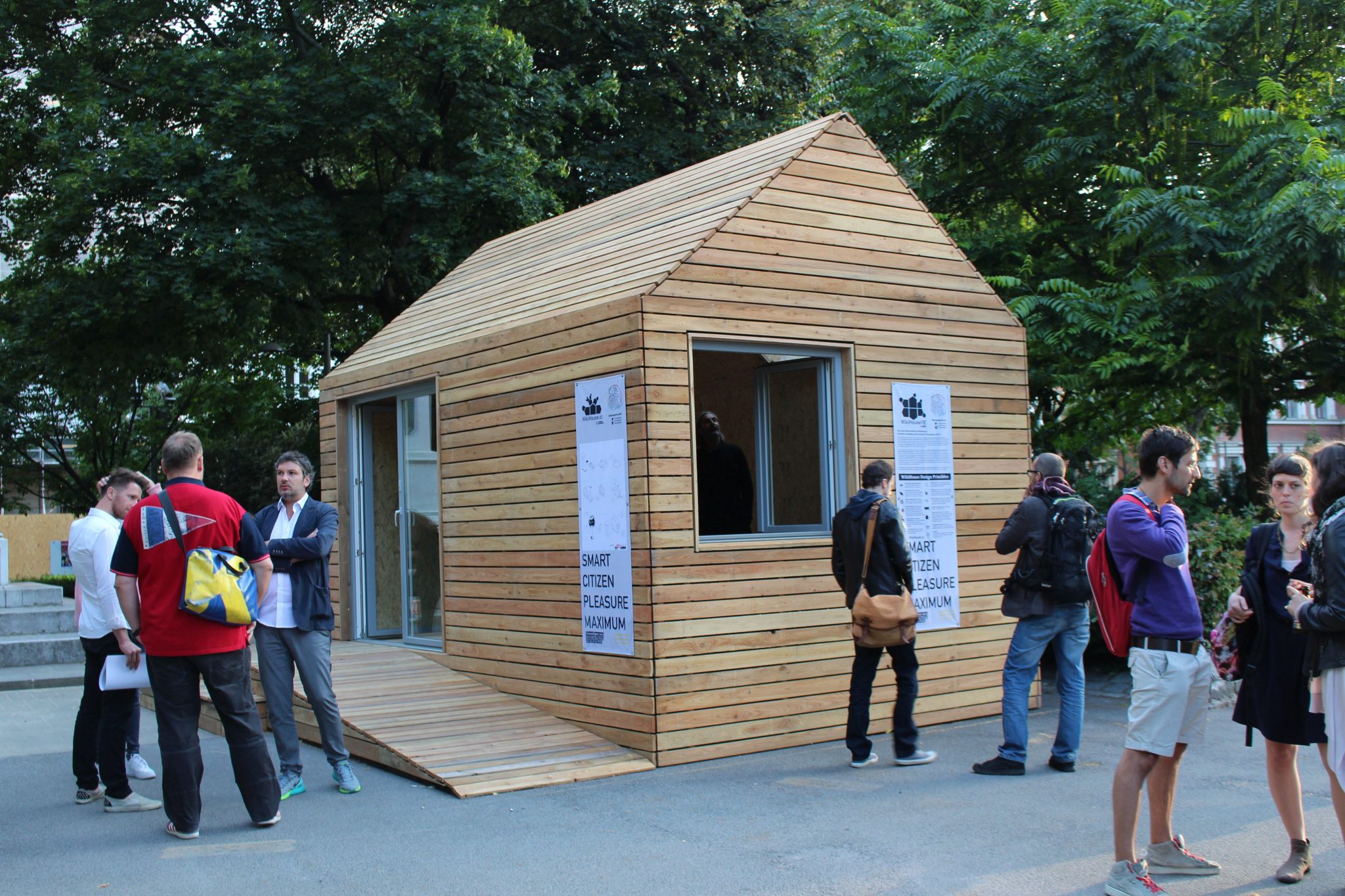 A small, modern wooden house with a sloped roof is surrounded by people engaged in conversation at an outdoor event. Two banners on the house read “SMART CITIZEN PLEASURE MAXIMUM.” Lush greenery and urban elements are in the background.