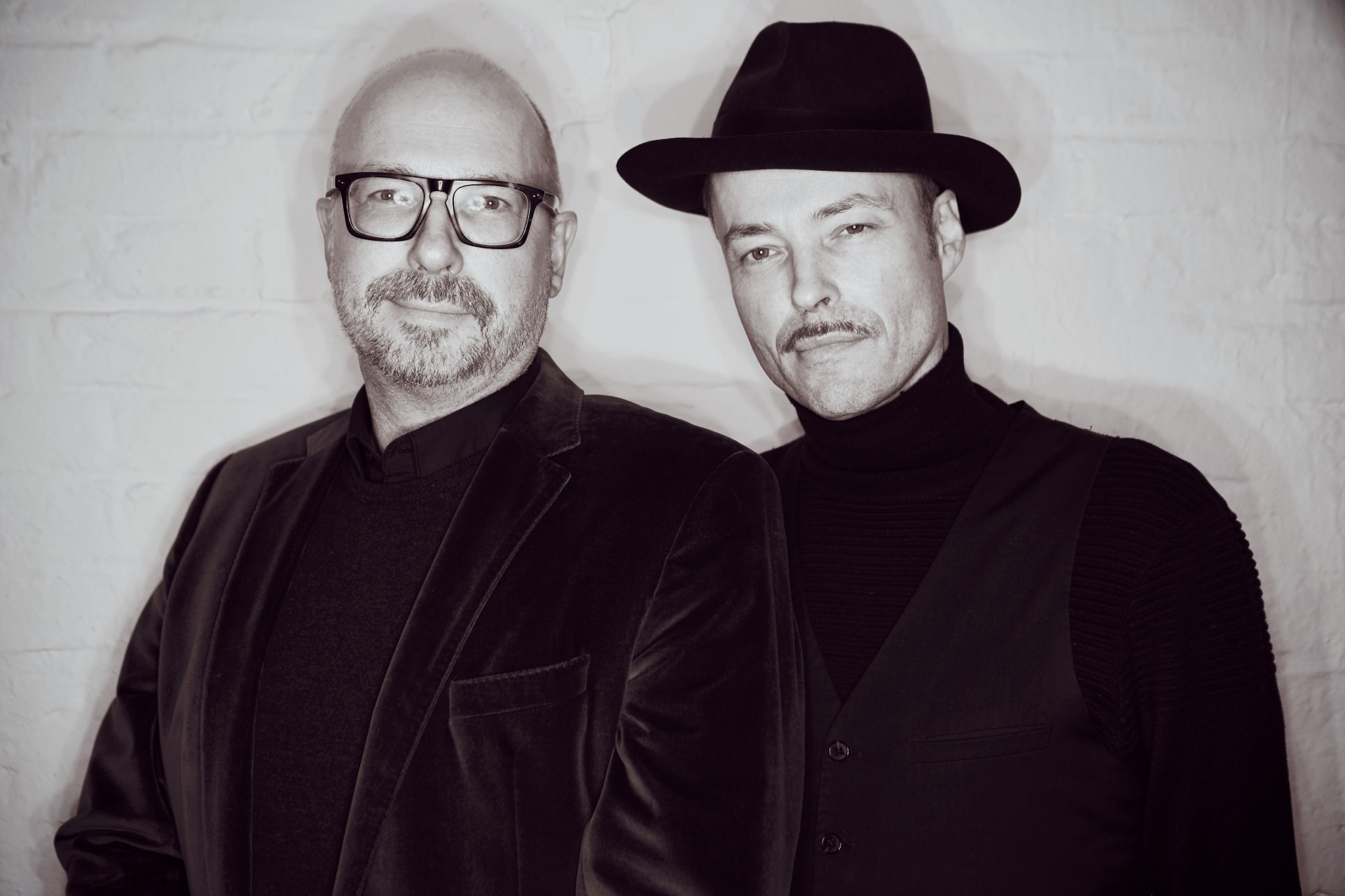 The image features three men standing in front of a white brick wall, with the central figure positioned slightly to the left. The man on the far right wears a black fedora hat and is attired in an all-black ensemble, comprising a turtleneck sweater and suit jacket. He also sports a thick mustache. The middle-aged individual to his left has shaved head and is dressed similarly, albeit without the hat and mustache. The third man, only partially visible on the far left, is wearing a black fedora hat and appears to be balding. The men's attire and facial hair suggest that they may be part of a band or musical group, possibly captured during a photo shoot.
