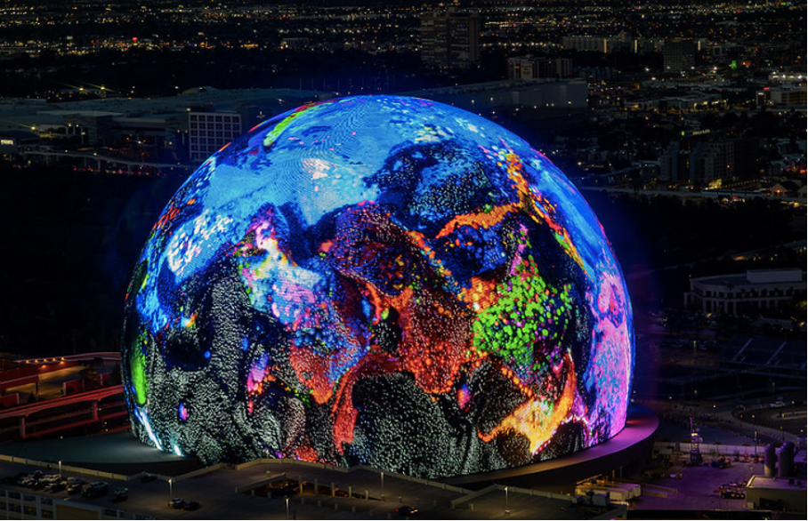 The image showcases the exterior of the Las Vegas Sphere, a large spherical structure illuminated at night. * The sphere is adorned with vibrant colors and patterns. * It has a large projection screen on its surface that displays various images and videos. * The lighting system creates a dynamic visual effect, changing colors and intensity throughout the day and night. * The sphere's exterior design features a unique blend of futuristic and artistic elements, making it a striking addition to the Las Vegas skyline. In summary, the image presents a visually stunning representation of the Las Vegas Sphere, highlighting its impressive design and technological capabilities.
