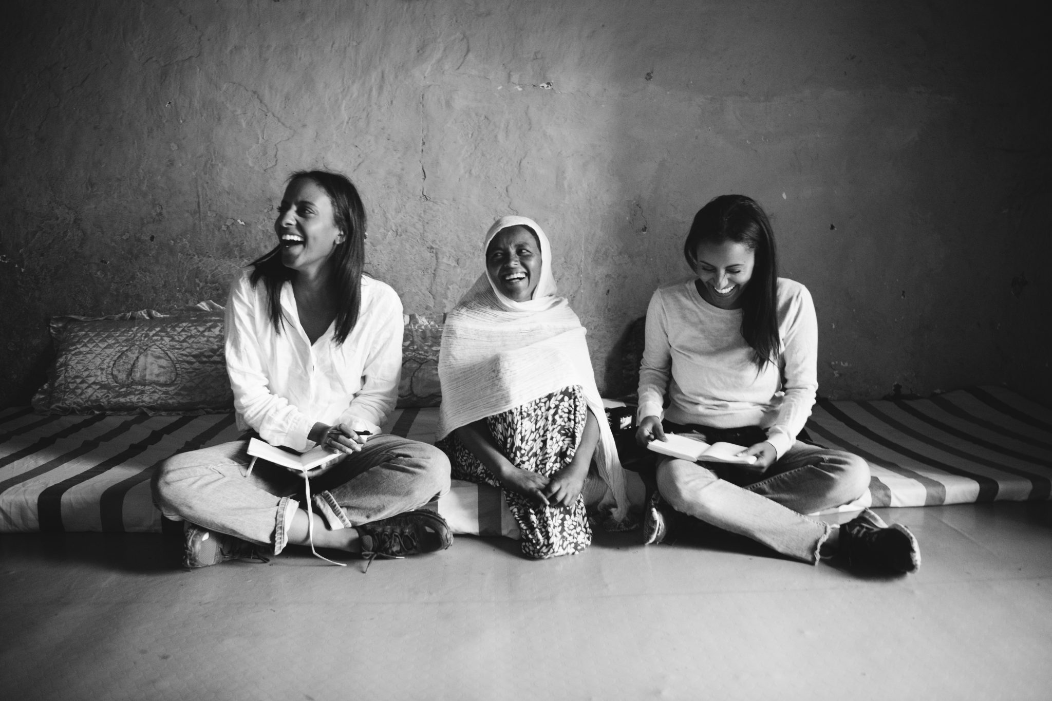 The image depicts three women sitting on the floor, engaged in various activities. In the foreground, a woman with long dark hair sits cross-legged, facing the camera. She wears a white shirt and jeans, holding a book or notebook in her lap. To her right is another woman, also seated cross-legged, wearing a headscarf and a patterned dress. The third woman, partially visible at the bottom of the image, has long dark hair and appears to be reading a book. The background features a wall with a rough texture, suggesting a rustic or natural setting. A striped mat or rug lies on the floor behind the women, adding a pop of color to the scene. Overall, the image conveys a sense of calmness and tranquility, as if the women are enjoying each other's company in a peaceful environment.