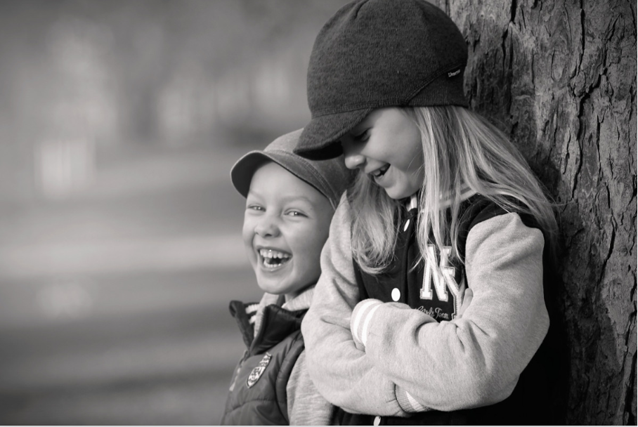 The image is a black-and-white photograph of two children playing together, with one child leaning against a tree trunk. * The boy in the middle has blond hair and wears a baseball cap, a dark jacket with a white fur collar, and a light-colored shirt underneath. He leans against a large tree trunk on his right side. * His facial expression is happy, and he appears to be laughing or smiling. * His left hand is raised near his shoulder, as if he is gesturing towards something off-camera. * The girl on the right also has blond hair and wears a dark jacket with white buttons down the front and a light-colored shirt underneath. She leans against the boy's back, resting her arms around him. * Her facial expression is also happy, and she appears to be laughing or smiling along with the boy. * The background of the image is out of focus, but it appears to be an outdoor setting, possibly a park or forest. Overall, the image captures a joyful moment between two children playing together in nature.