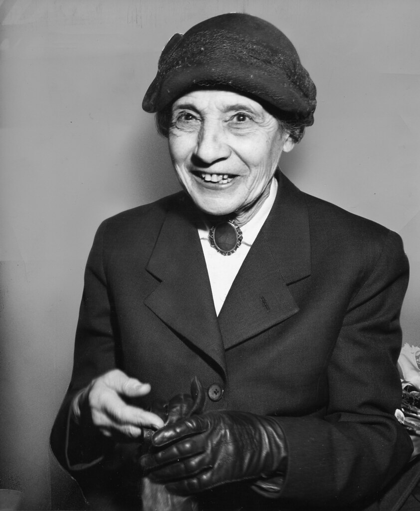 The image presents a black-and-white photograph of an elderly woman, likely taken in the mid-20th century, showcasing her dignified appearance. * **Woman's Clothing** * The woman is attired in a dark-colored coat with a single button at the top and lapels that extend to a point. * She wears a white collared shirt underneath, complemented by a brooch at the neckline featuring a large oval-shaped stone or gemstone. * Her head is adorned with a dark beret-like hat, possibly made of felt or wool, which adds a touch of elegance to her overall appearance. * **Accessories** * The woman's hands are elegantly gloved in leather gloves that extend up her arms, reaching just below the elbows. These gloves appear to be made from high-quality leather and have a smooth finish. * On her left hand, she wears a ring on her fourth finger, possibly signifying marriage or commitment. * **Background** * The background of the photograph features a light-colored wall, providing a subtle contrast to the woman's dark attire. In summary, the image captures a dignified elderly woman dressed in formal attire, complete with accessories that reflect her refined taste and sense of style.