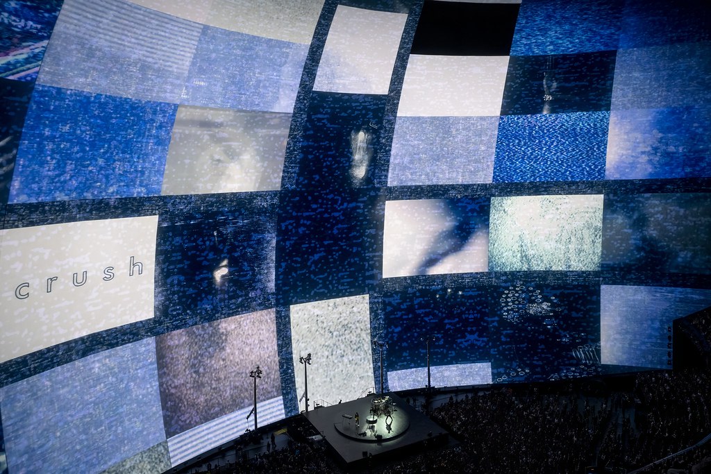 A large stage illuminated by a massive digital screen displaying various blue and textured visuals, with the word "crush" prominently featured; a small band performing in front of a captivated audience.