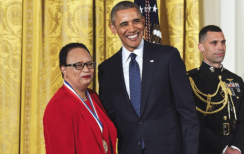 This image captures a moment of celebration at the White House, featuring Barack Obama presenting an award to Shirley Franklin, who is beaming with pride. In the foreground, Franklin stands out in her striking red suit jacket, adorned with a gold medal around her neck on a blue and white ribbon. She is accompanied by President Obama, dressed in his signature dark suit, white shirt, and blue tie. Notably, a small American flag pin is visible on his lapel. At the bottom of the image, an individual's arm and shoulder are partially visible, clad in a black military uniform with gold braiding and medals on the chest. The background features a yellow curtain with intricate designs, while the overall atmosphere exudes joy and celebration.