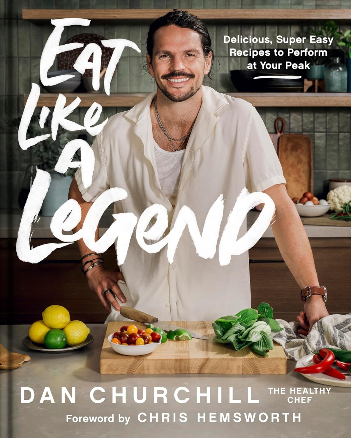 Cover of the cookbook "Eat Like a Legend" by Dan Churchill, featuring the author smiling while standing at a kitchen counter with fresh ingredients, including tomatoes, lemon, and greens, promoting delicious and easy recipes for peak performance.