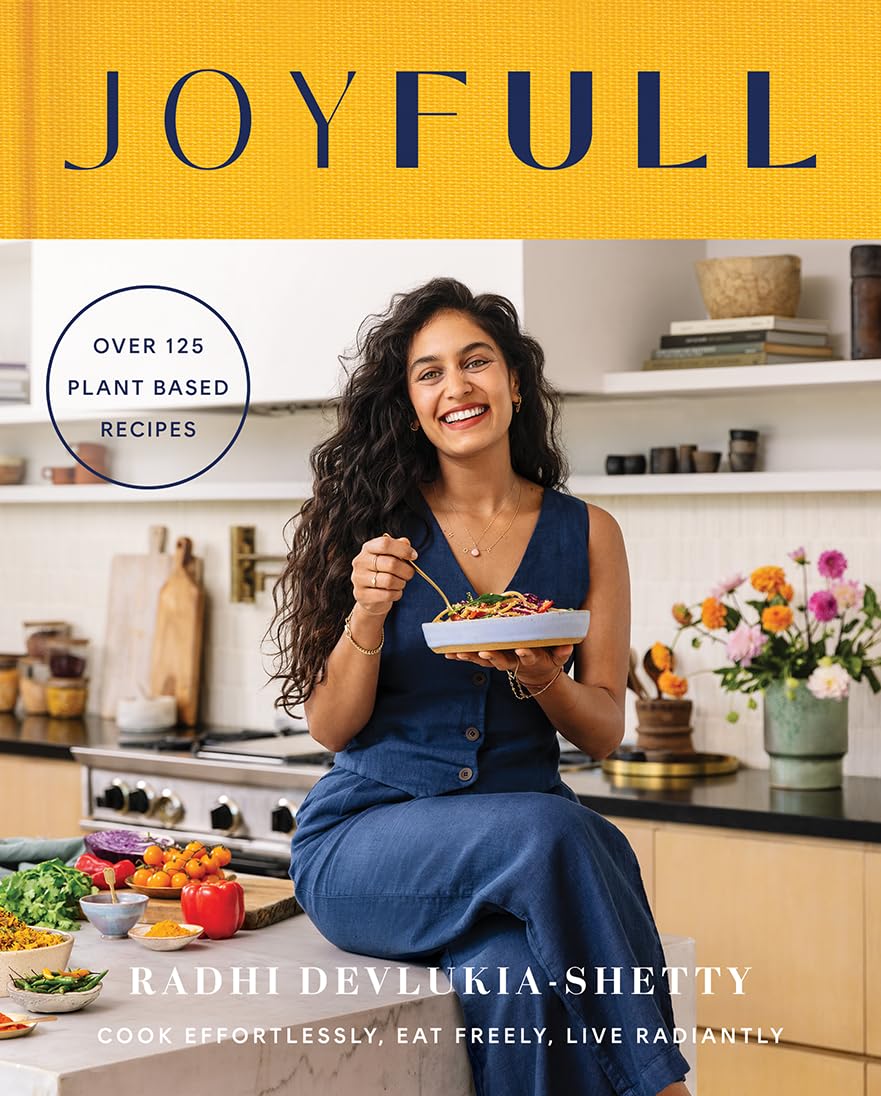 The image is a book cover for "JOYFULL" by Radhi Devlukia-Shetty, featuring a woman in a kitchen with various ingredients and cooking utensils. * The title of the book is "JOYFULL", written in large blue letters at the top of the image. + The word "JOYFULL" is centered on the cover, with the subtitle "OVER 125 PLANT BASED RECIPES" written below it in smaller text. * A woman with long dark hair and a denim dress is sitting on a counter, holding a plate of food. + She is wearing a denim dress with buttons down the front, and has a necklace around her neck. + The woman is smiling and looking at the camera, with a plate of food in front of her. * Various ingredients are scattered across the kitchen island. + There are vegetables such as bell peppers, tomatoes, and lettuce on the counter. + There are also bowls of nuts, seeds, and dried fruits visible. Overall, the image suggests that the book is about plant-based cooking and features a variety of healthy ingredients. The woman in the photo appears to be enjoying her meal, which adds to the overall sense of joy and satisfaction conveyed by the title "JOYFULL".