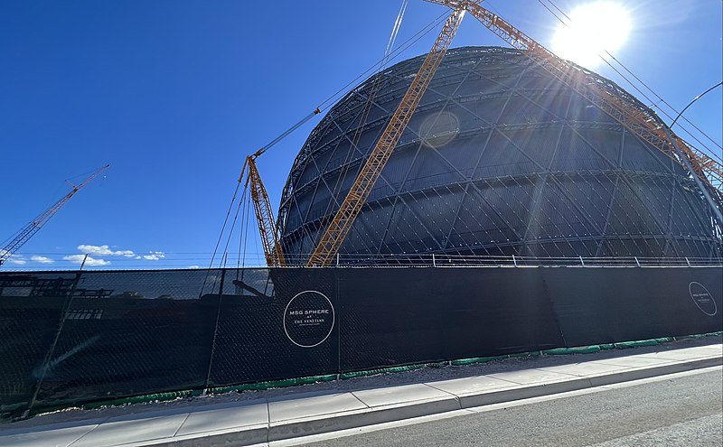 Construction site of a large spherical building, with cranes and fence in front, under a clear blue sky.