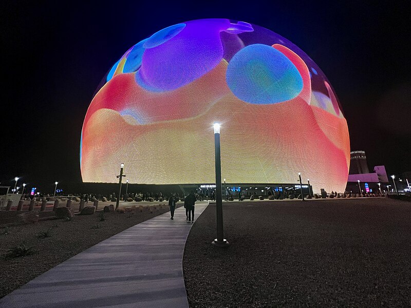 A brightly lit, large spherical structure displays colorful animated graphics at night, with people walking along a pathway in the foreground.