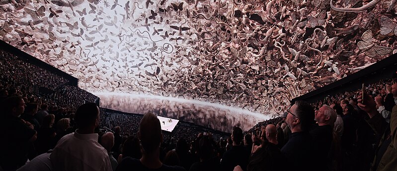 This image captures the interior of an art gallery or museum, where a large crowd has gathered to view a striking painting on the ceiling. The artwork features a predominantly white background adorned with pinkish-red designs that resemble paint splatters or abstract shapes. The surrounding walls and floor are shrouded in darkness, creating a dramatic contrast between the vibrant ceiling piece and the dimly lit space. In the background, a few individuals can be seen standing near the wall, while others are positioned closer to the artwork. The overall atmosphere is one of awe and fascination, as visitors marvel at the unique and captivating installation on display.