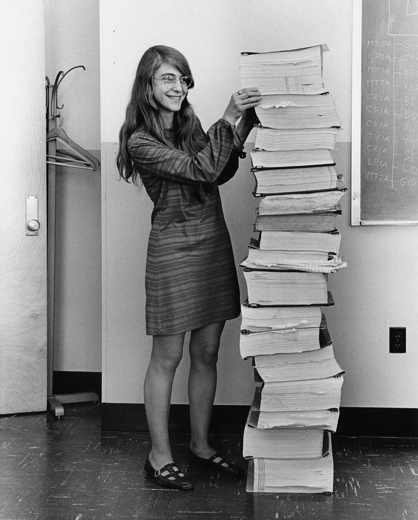 The image depicts a woman standing beside a tall stack of papers, likely in an office setting. In the foreground, the woman is positioned on the left side of the image, facing right. She has long dark hair and wears a short dress with stripes, paired with black shoes. Her hands are raised to hold up the topmost paper in the stack, which appears to be slightly bent. The stack itself consists of numerous papers, some folded or rolled up, with no discernible order or organization. In the background, a white wall provides a neutral backdrop for the scene. A coat rack is visible on the left side of the image, while a chalkboard or whiteboard hangs on the right-hand side, displaying some illegible writing in white chalk. The overall atmosphere suggests a busy office environment, possibly during a period of intense paperwork or document management. The woman's pose and expression convey a sense of pride or accomplishment, as if she has successfully managed to stack the papers in this manner. Her attire and hairstyle suggest that she may be from an earlier era, perhaps the 1960s or 1970s. The image provides insight into the daily work life of individuals during this time period, highlighting the importance of record-keeping and document management in various industries.
