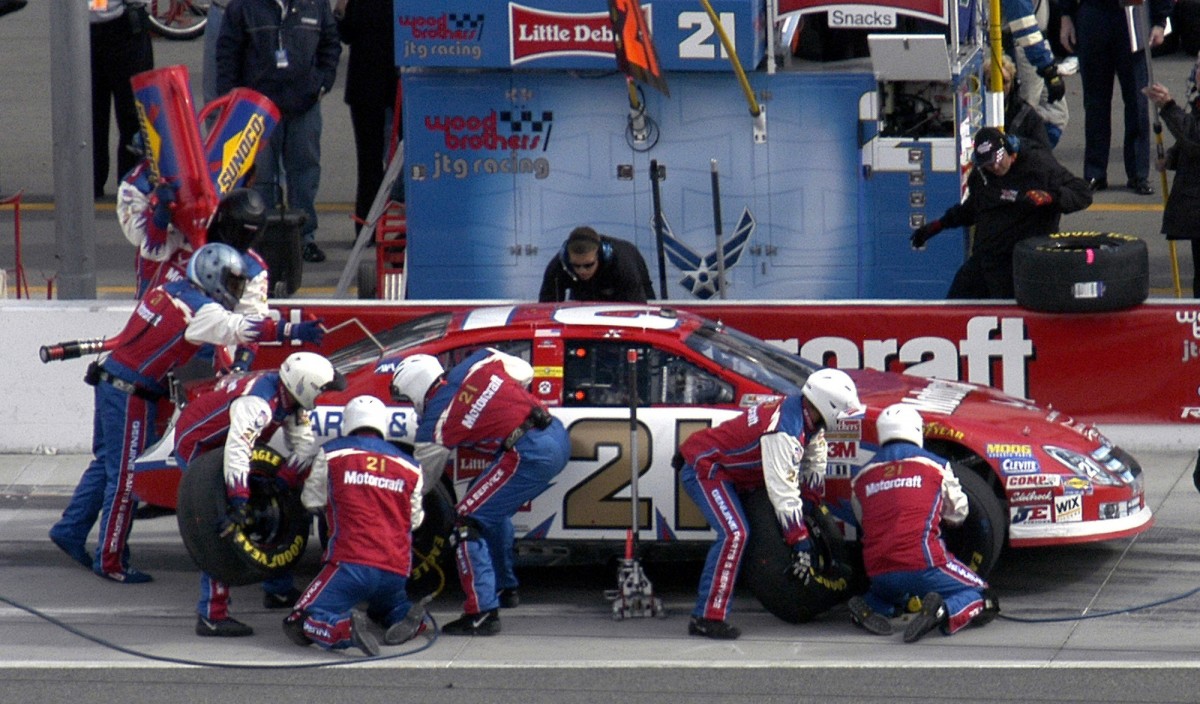 The image depicts a NASCAR pit crew working on a race car during a pit stop. * The crew is dressed in matching uniforms with red, white, and blue colors. * They are wearing helmets to protect themselves from debris and other hazards. * Each member has a specific role, such as fueling, tire changing, or adjusting the car's settings. * The race car is parked on the pit lane, with its hood up and tires exposed for maintenance. * The crew is busy working together to complete the pit stop efficiently. * They are using specialized tools and equipment to perform their tasks quickly and safely. * In the background, spectators can be seen watching the pit stop from behind a fence or barrier. * They appear to be excited and engaged in the action unfolding before them. * The overall atmosphere suggests a high level of competition and intensity, as each team strives to gain an advantage over their rivals. Overall, the image captures the fast-paced and dynamic nature of NASCAR pit stops, highlighting the importance of teamwork, strategy, and quick thinking in achieving success on the track.