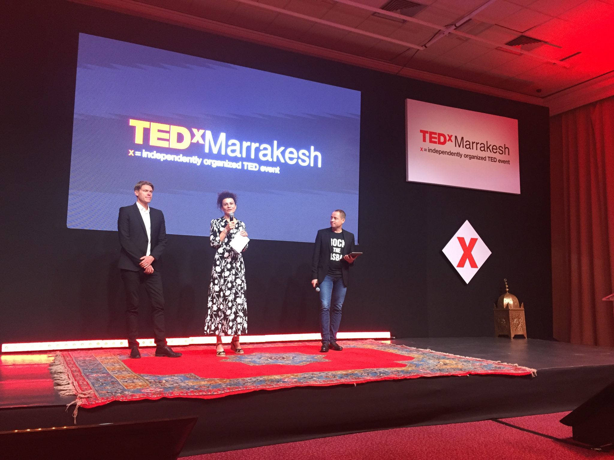 Three presenters on stage at the TEDxMarrakesh event, with a large screen displaying the event title, as they engage with the audience. The stage is adorned with a colorful rug and decorative elements.
