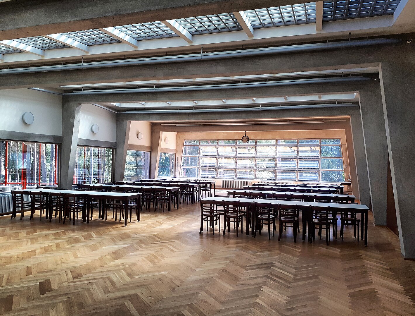 The image showcases a spacious, well-lit room with a polished wooden floor featuring an intricate parquet design. The room is furnished with rows of long tables and chairs, creating a sense of orderliness. **Key Features:** * **Skylights:** Natural light pours in through the skylight windows on the ceiling, illuminating the room. * **Large Windows:** The walls are adorned with large windows that provide an abundance of natural light, while also offering a view of the surrounding area. * **Polished Wooden Floor:** The floor is made of polished wood and features a parquet design, adding warmth and sophistication to the space. **Overall Impression:** The image presents a clean, well-maintained room that exudes a sense of professionalism. The abundance of natural light and the polished wooden floor create a welcoming atmosphere, making it an ideal setting for various purposes such as meetings, events, or everyday activities.