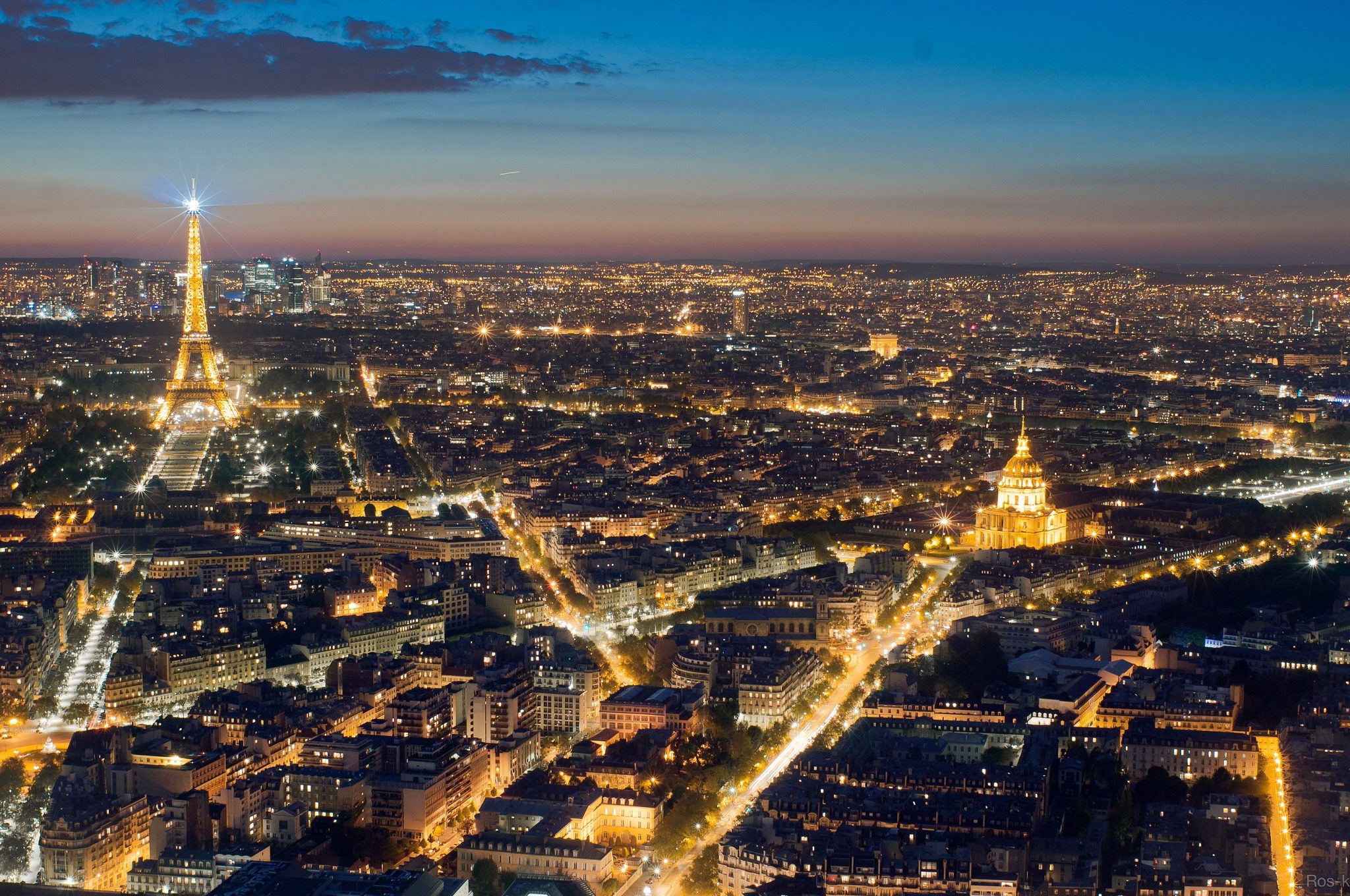 Aerial view of Paris at dusk, featuring the illuminated Eiffel Tower on the left and the golden Dome des Invalides on the right, with the city lights twinkling below against a twilight sky.