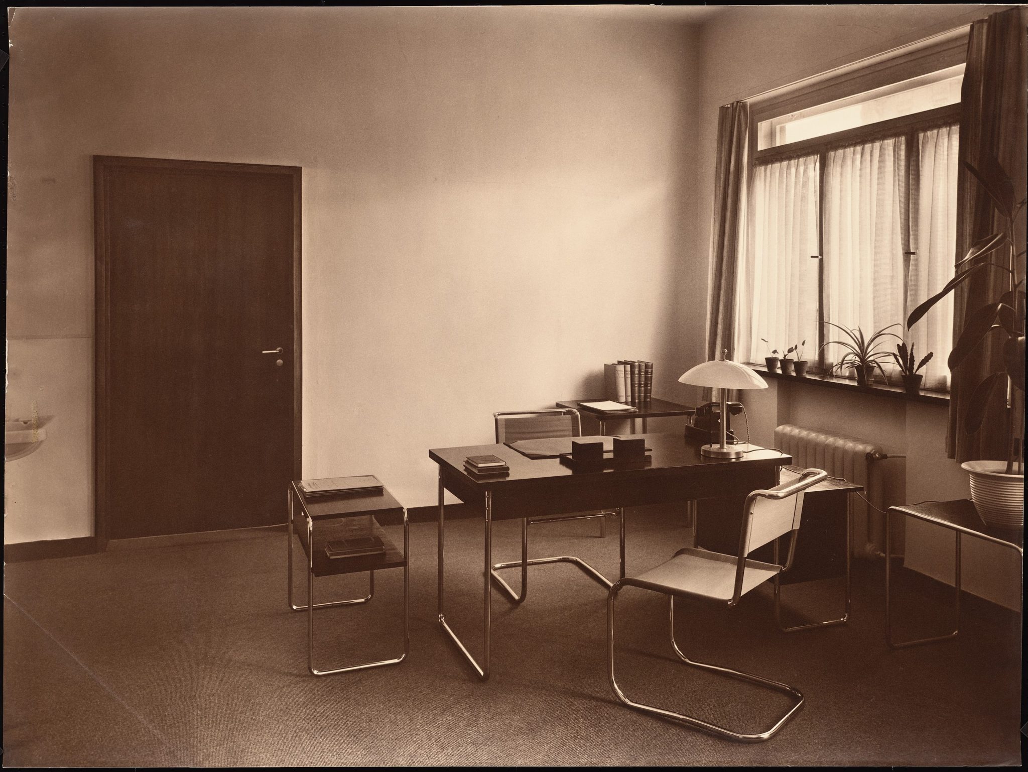Minimalist office interior featuring a wooden desk, two chairs, a lamp, and a window with sheer curtains, complemented by potted plants and a closed door.
