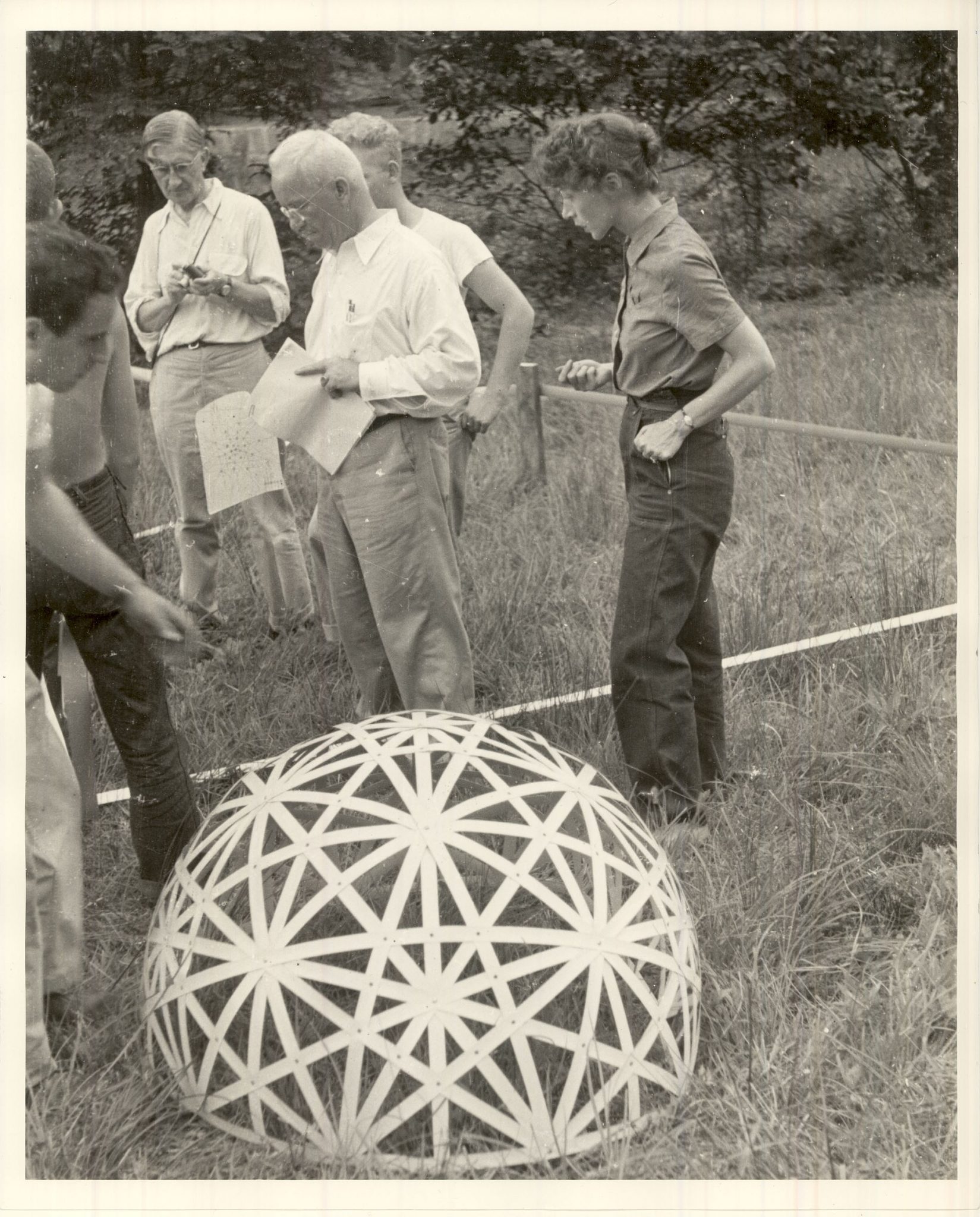 The image depicts a group of people gathered around an intricately designed, spherical object in a grassy field. The black-and-white photograph captures the scene with clarity. In the foreground, a large, round sphere dominates the center of the frame. Its surface is adorned with a pattern of intersecting lines that form a geometric design, evoking the appearance of a dodecahedron. The sphere sits on a grassy field, surrounded by tall blades of grass. Behind the sphere, several individuals are visible, standing in a relaxed manner. They appear to be engaged in conversation or observing the sphere's intricate pattern. One person is holding papers, suggesting they may be discussing or studying the design. The background of the image features trees and other foliage, providing context for the outdoor setting. Overall, the photograph presents a captivating scene that invites viewers to ponder the significance and purpose of the spherical object and its geometric pattern.