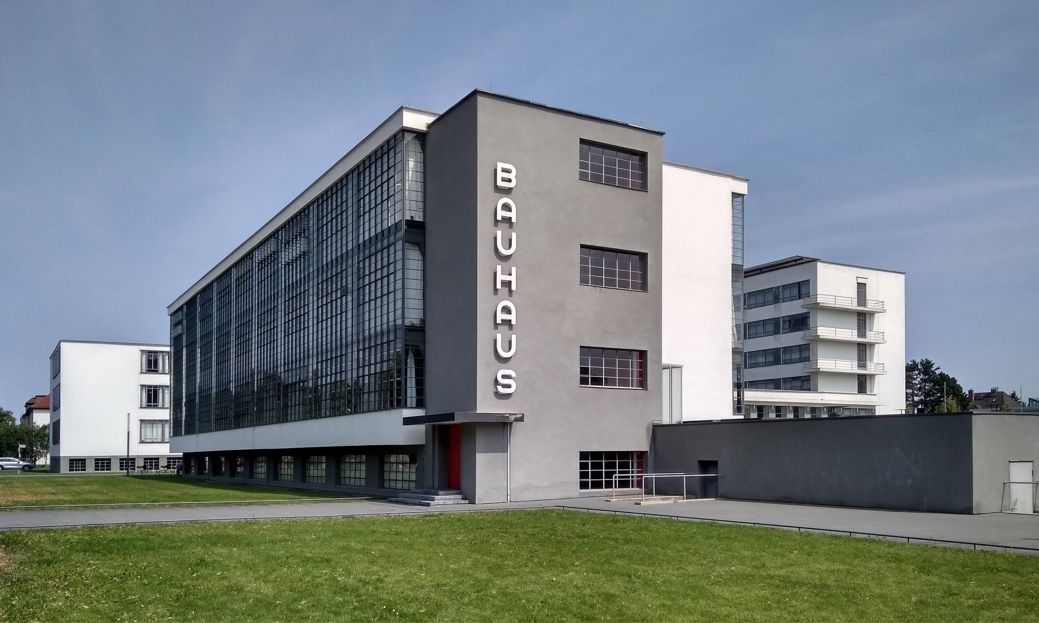 The image depicts a large, multi-story building with the word "BAUHAUS" written vertically on its side in white letters. * The building is situated in an open area, possibly a campus or park. * It has multiple stories and a flat roof. * The windows are rectangular and arranged in rows. * The entrance to the building is located at the bottom of the structure. * A large sign reading "BAUHAUS" is displayed on the side of the building. The overall atmosphere suggests that this is a school or educational institution, given the presence of multiple stories and classrooms.