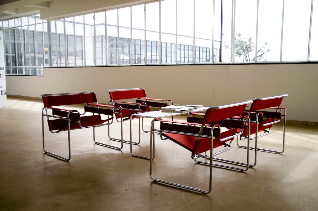 This photograph showcases an open-plan office space with a striking red chair arrangement, featuring four chairs positioned around a table on the right side of the room. The chairs boast sleek chrome frames and rich red leather upholstery, while the table's surface is cluttered with papers or books. The background features a wall adorned with floor-to-ceiling windows that allow an abundance of natural light to pour in, casting a warm glow over the space.