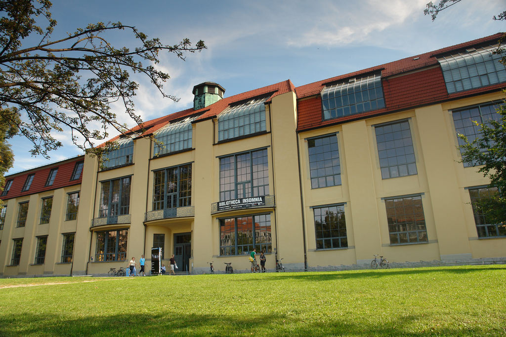 A modern library building with large windows and a distinctive architectural style, set on a grassy area with people and bicycles in view. The sign reads "Biblioteca Insomnia."