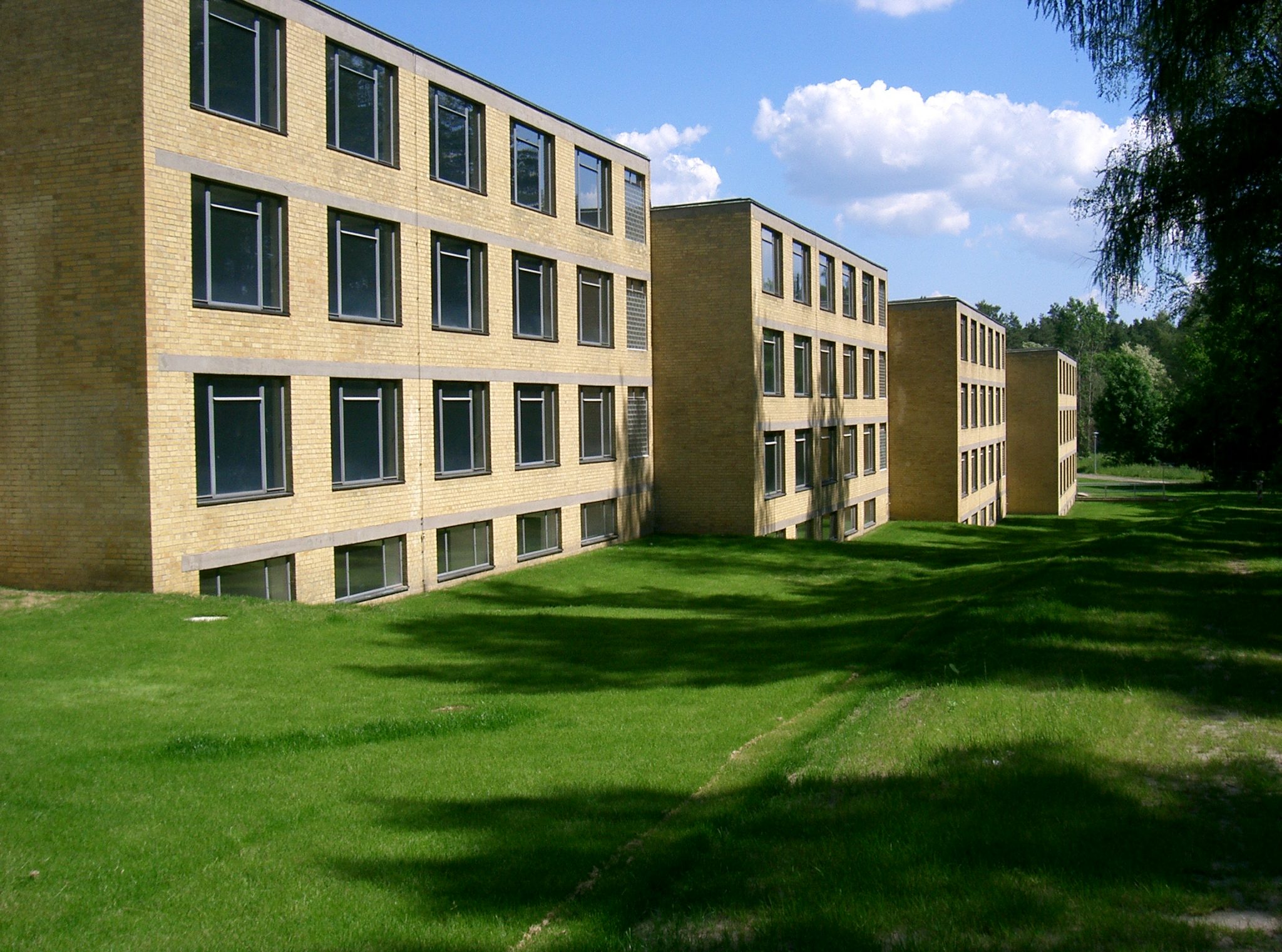 The image depicts three tan brick buildings with numerous windows situated on a lush green lawn, evoking an atmosphere reminiscent of a university or college campus. In the foreground, three identical buildings are visible, each featuring multiple windows on their front sides. The buildings' façades appear to be constructed from tan bricks and have flat roofs. They are positioned on a well-manicured grassy area, with trees visible in the background. The sky above is blue and cloudy, suggesting that the photo was taken during the daytime. The image exudes a sense of serenity and tranquility, as if it were captured on a peaceful university campus.