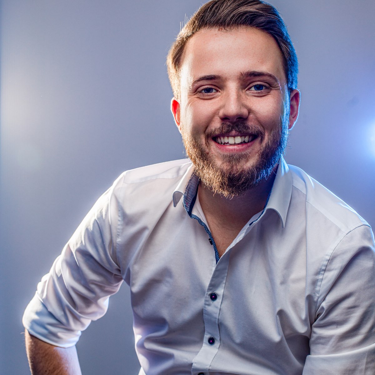 Smiling young man with a beard wearing a white shirt, posing against a gradient background with soft lighting.