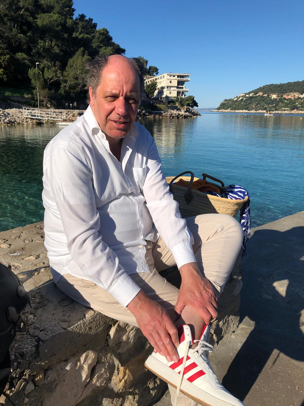 A man in a white shirt and beige pants sits on a stone pier by a calm waterfront, tying his red and white sneakers, with trees and a hillside in the background.