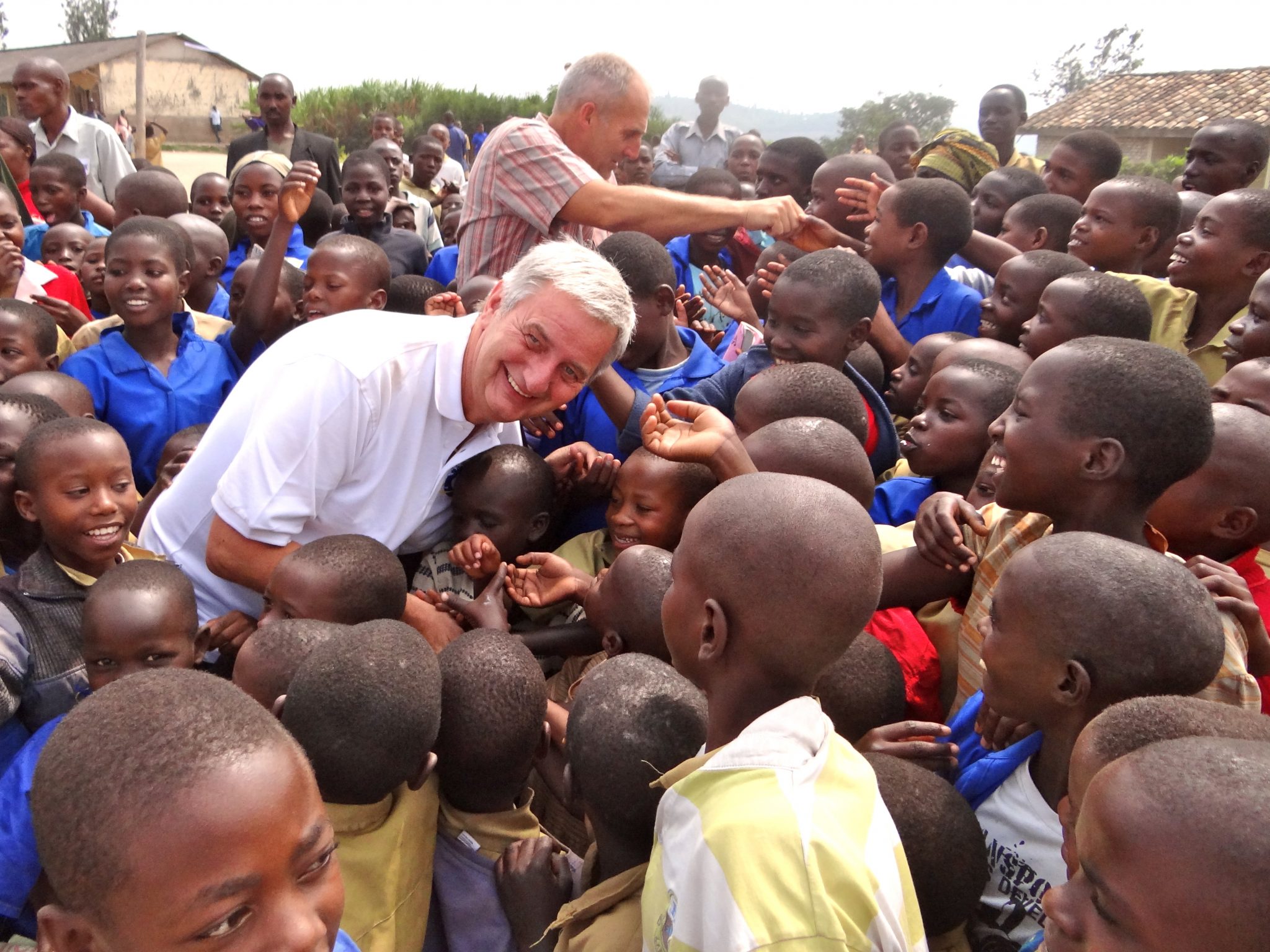 The image depicts a heartwarming scene of an older man surrounded by numerous children, all gathered around him with their faces filled with joy and admiration. In the foreground, the man, dressed in a white polo shirt, is seen leaning down to engage with the children. The majority of them are wearing blue shirts, while some wear yellow or red tops. Their hands are extended towards the man, conveying a sense of warmth and connection. Behind the crowd, two men can be seen standing near the top left corner of the image. One of them is reaching out with his right arm to touch the head of one of the children. The group appears to be situated outside a building that resembles a school or church, set against an overcast sky. The overall atmosphere of the image exudes warmth and kindness, as the older man's gentle demeanor and the children's enthusiastic reception create a sense of harmony and mutual respect.