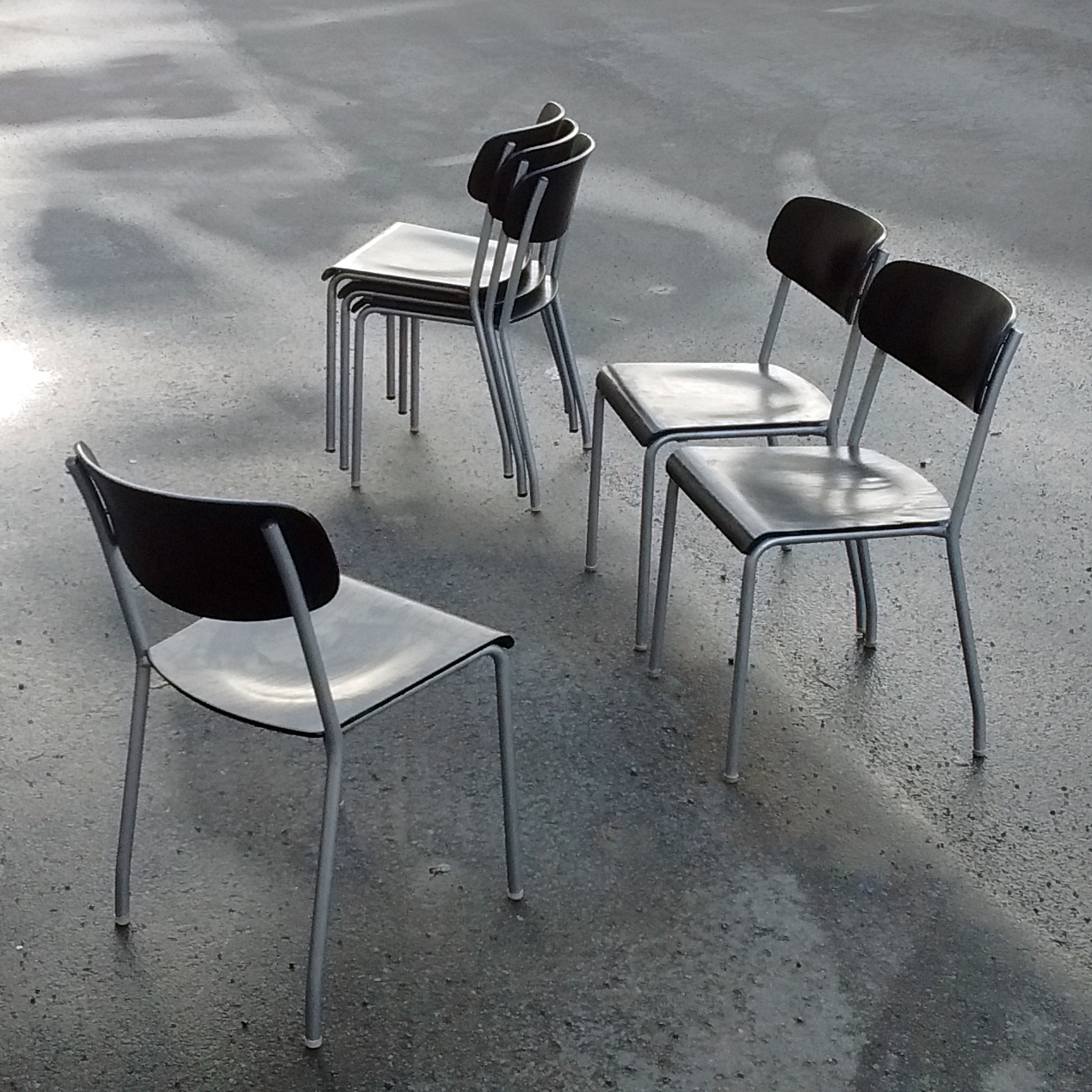 This photograph features four metal chairs situated on a concrete floor. The chairs are arranged in two rows of two, with the front row positioned closer to the camera and slightly angled away from it. The back row is placed at an angle facing left, creating a sense of depth and perspective within the image. Each chair boasts a sleek silver-colored frame, complemented by black plastic seat and backrests that appear to be made of a shiny material, possibly vinyl or faux leather. The chairs' minimalist design allows their individual features to shine through, while the concrete floor provides a stark contrast in texture and tone. The overall mood of the image is one of simplicity and functionality, with the chairs appearing to be in good condition and ready for use. The absence of any other objects or distractions in the background emphasizes the focus on the chairs themselves, creating a sense of clarity and directness that draws the viewer's attention.