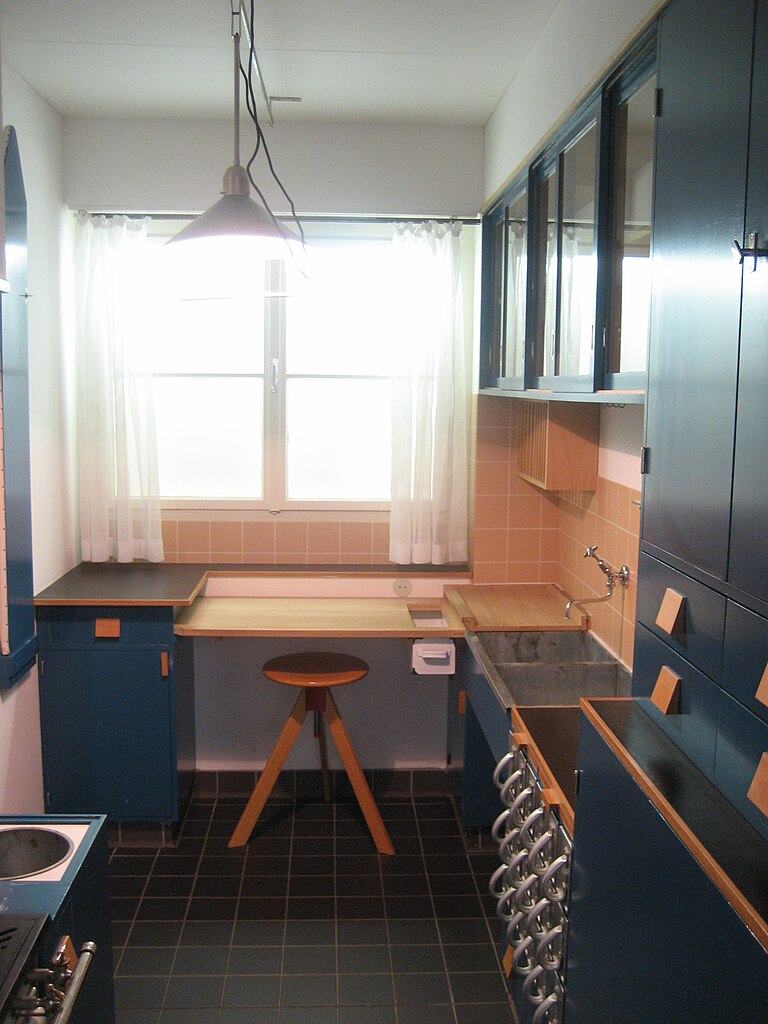 This image depicts a compact kitchen with black cabinets, tile flooring, and white walls. In the foreground, on the left side of the room, is an L-shaped counter with a dark grey countertop and matching cupboards underneath. The back wall features two windows with sheer curtains in front of them. Above one window, there are three cabinet doors that open outwards, each containing a silver handle. Below this row of cabinets, another countertop spans the width of the room, providing space for food preparation or cooking. On the right side of the image, an oven is visible at the bottom of the frame, with its door closed and a white knob on top. Above the oven are more cabinets, including two drawers that open outwards from the wall in front of them. The tile flooring provides a cohesive look throughout the room, tying together the various elements. Overall, this image showcases a functional and stylish kitchen design, with a focus on simplicity and practicality.