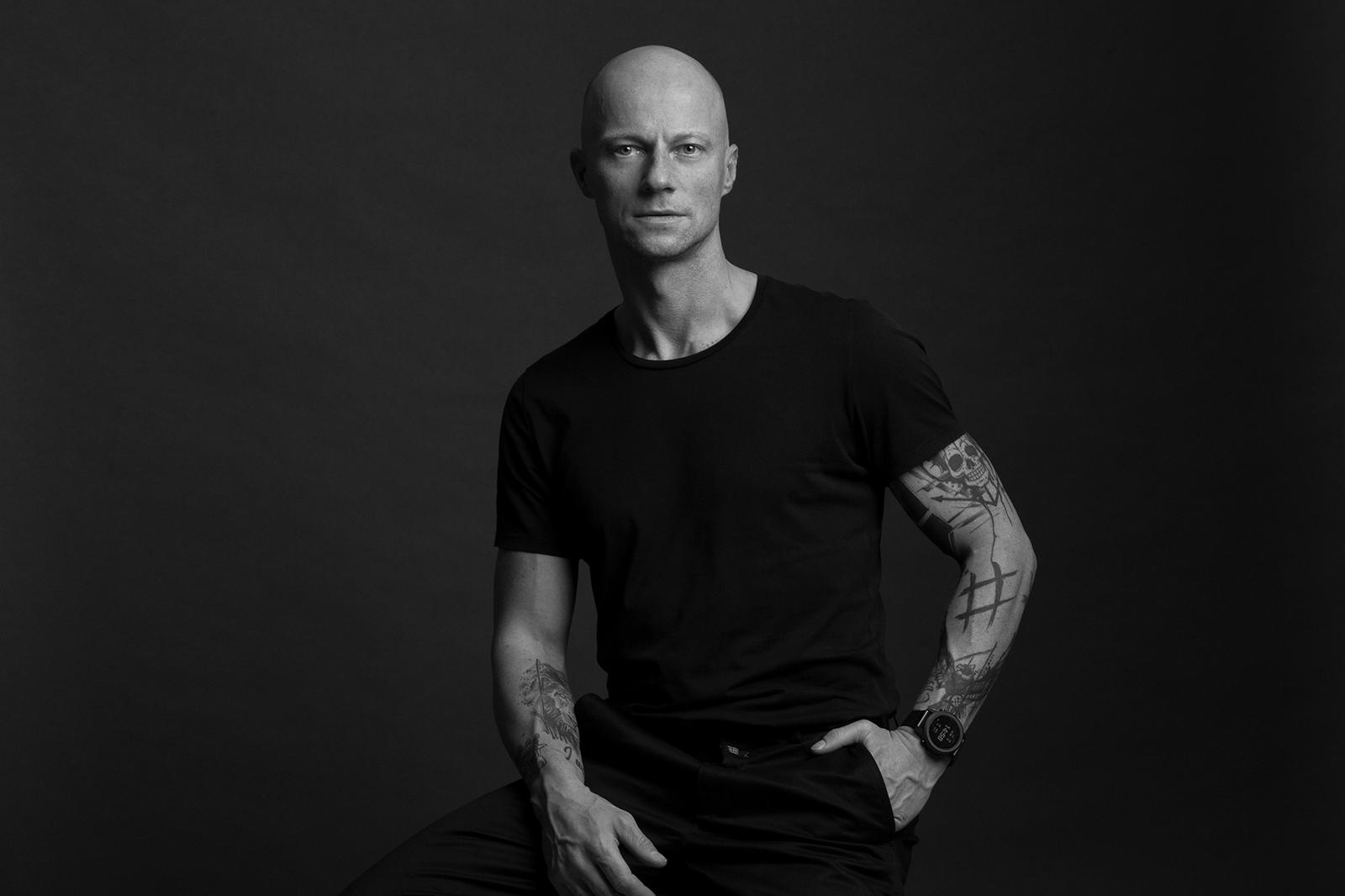 A bald man with tattoos is sitting confidently against a dark background, wearing a black t-shirt and black pants.