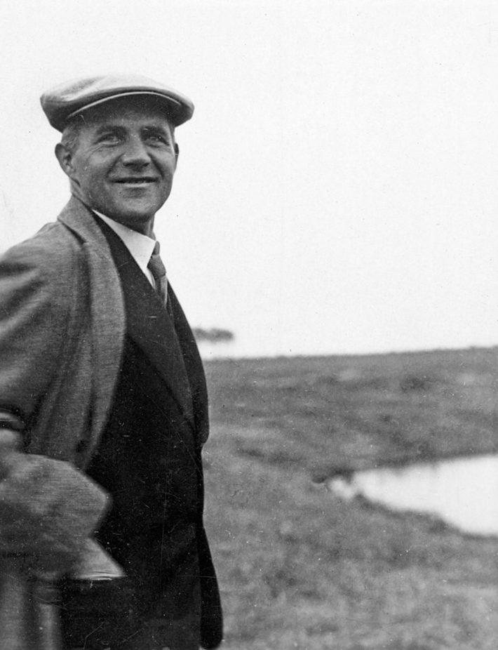 The image presents a black-and-white photograph of an older gentleman donning a flat cap, standing in a field with a serene body of water in the background. In the foreground, the man is attired in a dark suit, complemented by a crisp white collared shirt and a tie, which adds a touch of elegance to his overall appearance. The flat cap, a classic accessory of its time, sits atop his head, completing his dignified look. His gaze is directed towards the camera, exuding a sense of confidence and poise. As he stands in the field, the man's presence is juxtaposed with the tranquil atmosphere of the surrounding landscape. The body of water in the background serves as a subtle reminder of the natural beauty that exists beyond the confines of human activity.