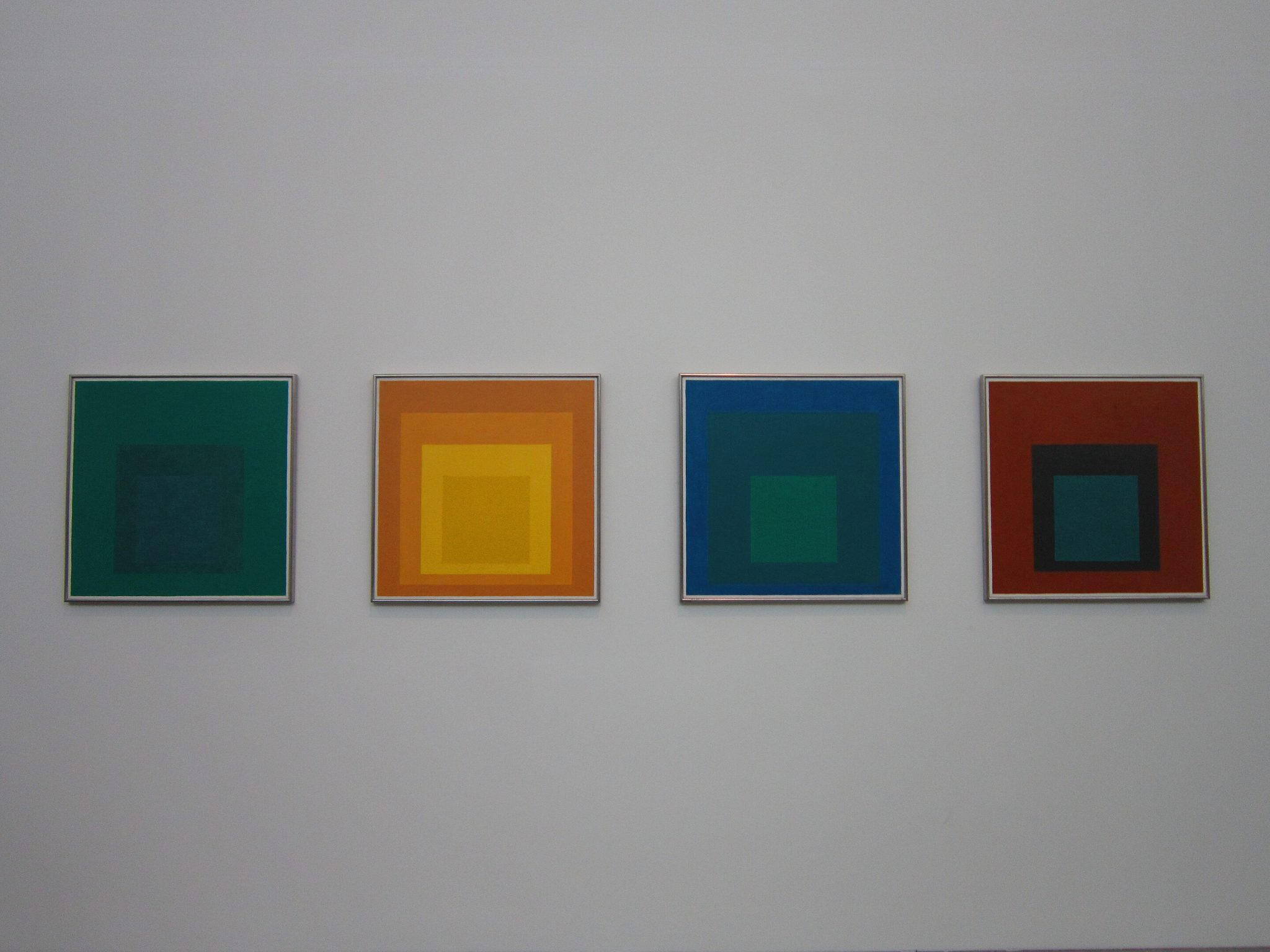 The image presents a series of four square paintings, each with a distinct color scheme and design. * The first painting features a dark green background. + The background is solid, without any additional details or patterns. + It provides a subtle yet rich tone to the overall composition. * The second painting boasts a vibrant orange hue as its background. + A lighter shade of orange forms a square in the center of the canvas. + This creates a sense of depth and visual interest within the piece. * The third painting showcases a deep blue color for its background. + Similar to the previous one, a lighter shade of blue is centered on the canvas. + This design element adds complexity and intrigue to the artwork. * The fourth painting has a reddish-brown background. + A smaller square in a darker shade of green is situated at the center of the composition. + This creates a sense of balance and harmony within the piece. In summary, each painting in this series employs a distinct color palette while maintaining a consistent design element. The use of contrasting colors adds visual appeal and depth to the overall image.
