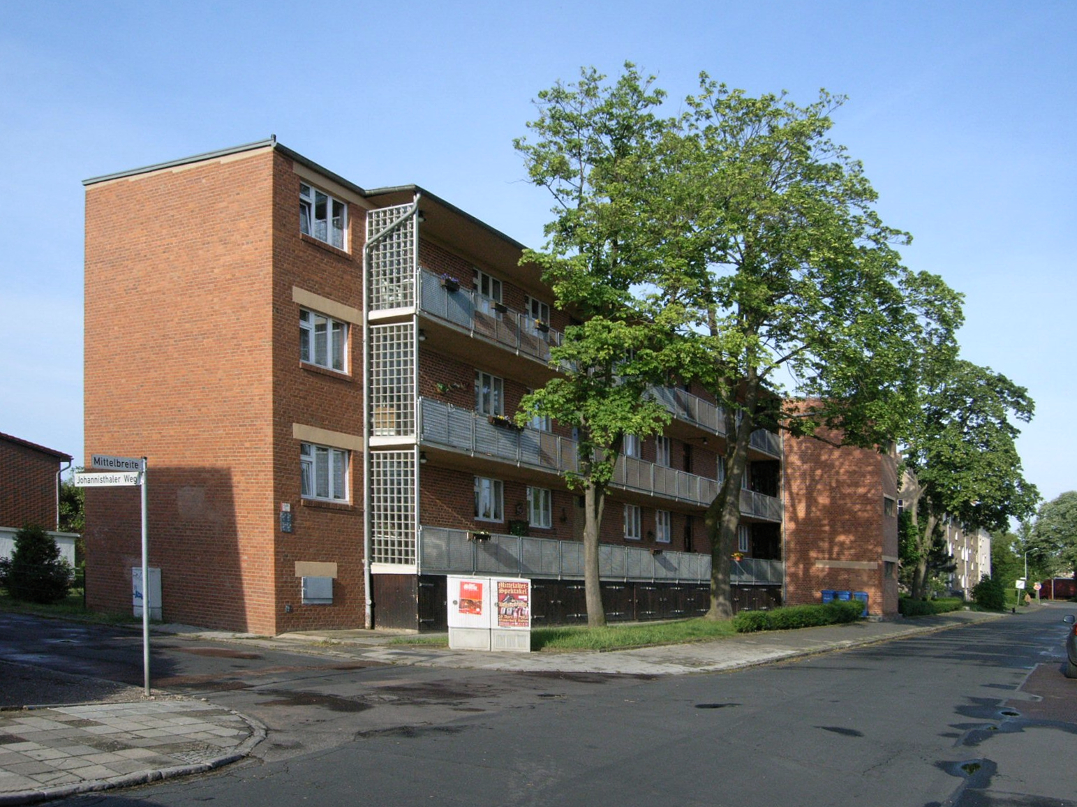 The image depicts a multi-story apartment building situated on the corner of a street, with a tree in front of it and a blue sky above. * The building is made of brick and has multiple floors. + Each floor appears to have several windows. + There are also some balconies visible on the upper floors. * A large tree stands in front of the building, partially obscuring its view. + The tree's branches stretch out towards the street. + Its leaves are a vibrant green color. * The sky above is a clear blue. + No clouds or other obstructions are visible. + The sun appears to be shining down on the scene. The image presents a serene and peaceful atmosphere, with the brick building and tree creating a sense of stability and growth.