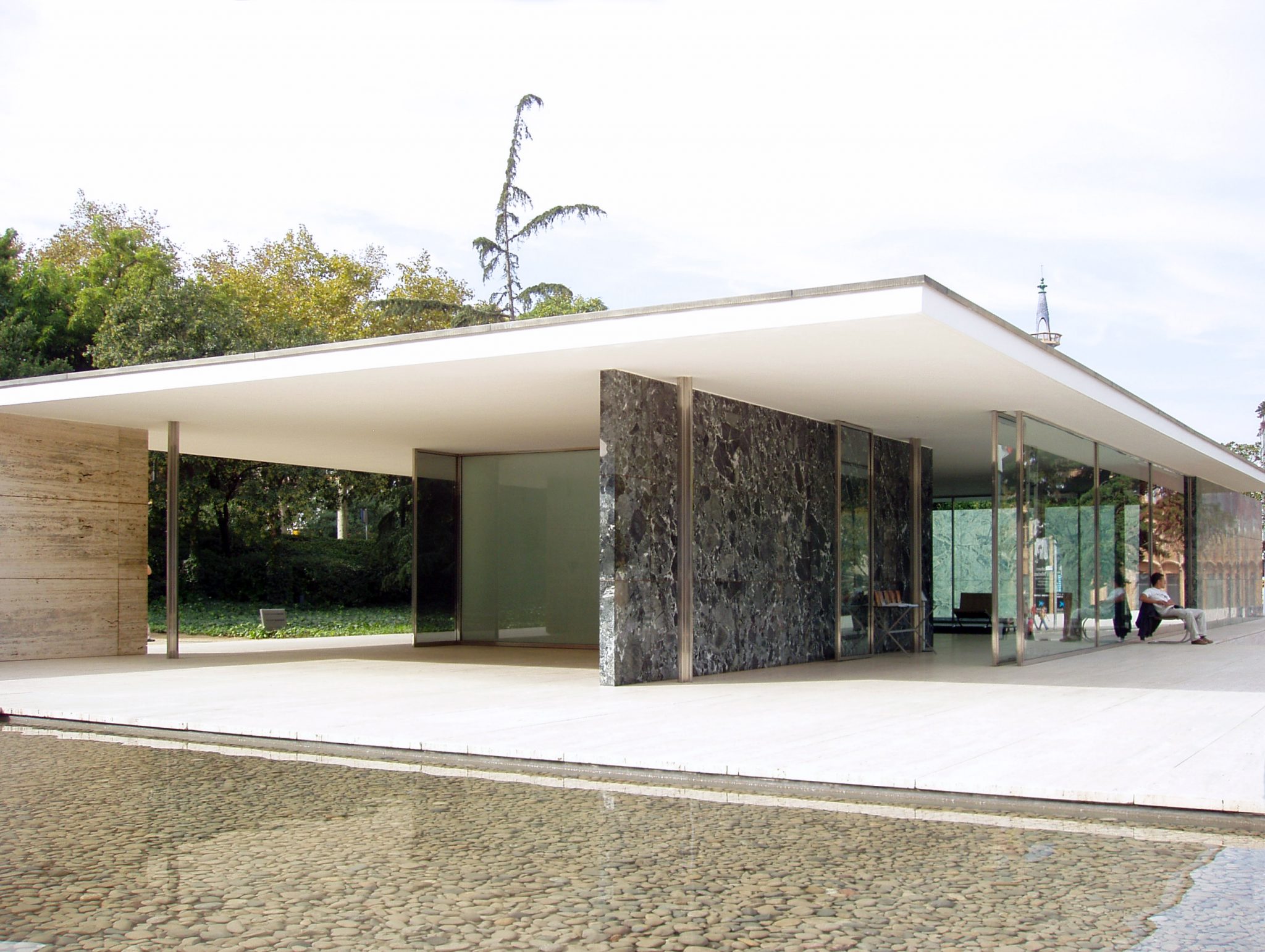 This photograph captures the exterior of the Barcelona Pavilion, a renowned building designed by Ludwig Mies van der Rohe for the 1929 International Exposition in Barcelona, Spain. The pavilion's modernist design features a striking white roof that extends over its front and right sides, supported by slender metal columns. The left side is made entirely of glass and steel, allowing natural light to flood the interior. The surrounding landscape includes lush greenery, while visitors are visible on the right side of the building. The image conveys a sense of elegance and sophistication, showcasing the pavilion's enduring appeal as a cultural icon of modern architecture.