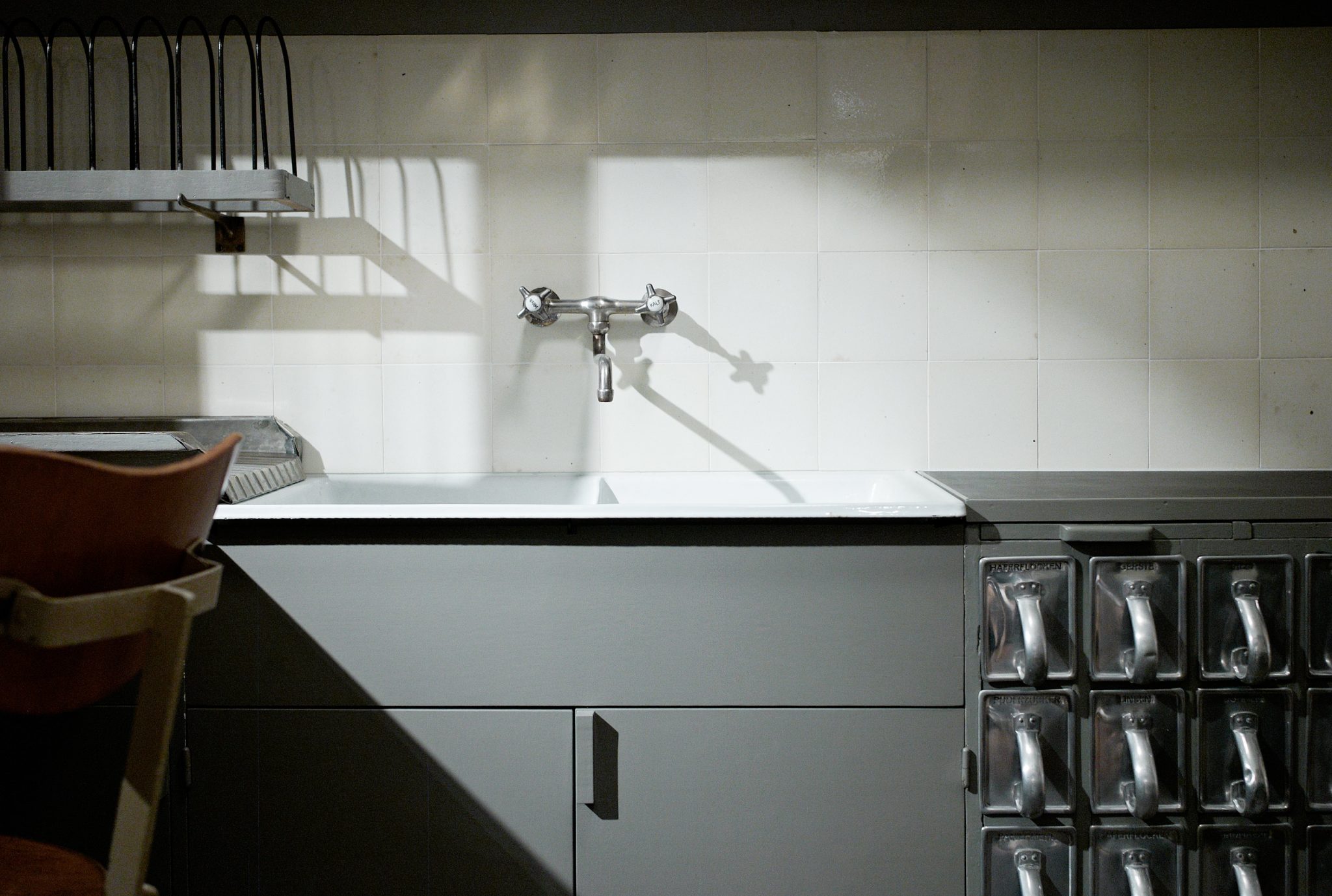 Alt tag: A modern kitchen sink with a metallic faucet against a tiled wall, featuring a metal dish rack above and a cabinet with metal drawers on the right. A wooden chair is partially visible on the left.