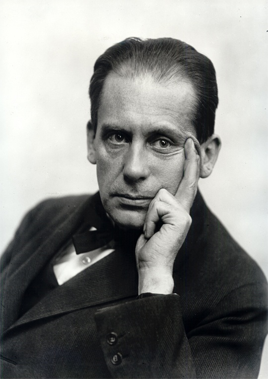 Black and white portrait of a man in formal attire, resting his chin on his hand with a contemplative expression.