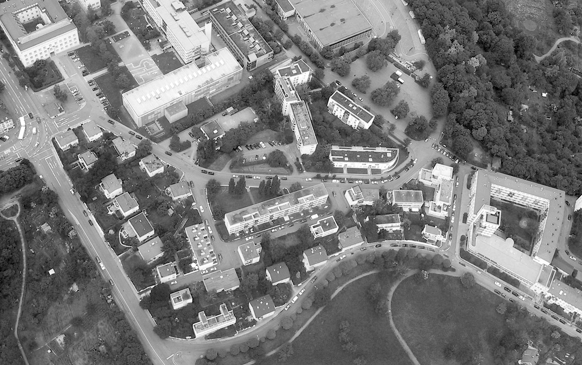 This aerial photograph presents an urban landscape in grayscale, showcasing the layout of buildings and roads within a specific area. The image features two main roads that intersect at its center, with one running horizontally across the top portion of the photo and another curving through the lower half, creating a sharp right angle. The surrounding streets are lined with trees, while several multi-story buildings with flat roofs dominate the scene. One building stands out for its distinctive pointed roof. The image's grayscale tone suggests it was taken on an overcast day or edited to emphasize structure and detail over color.