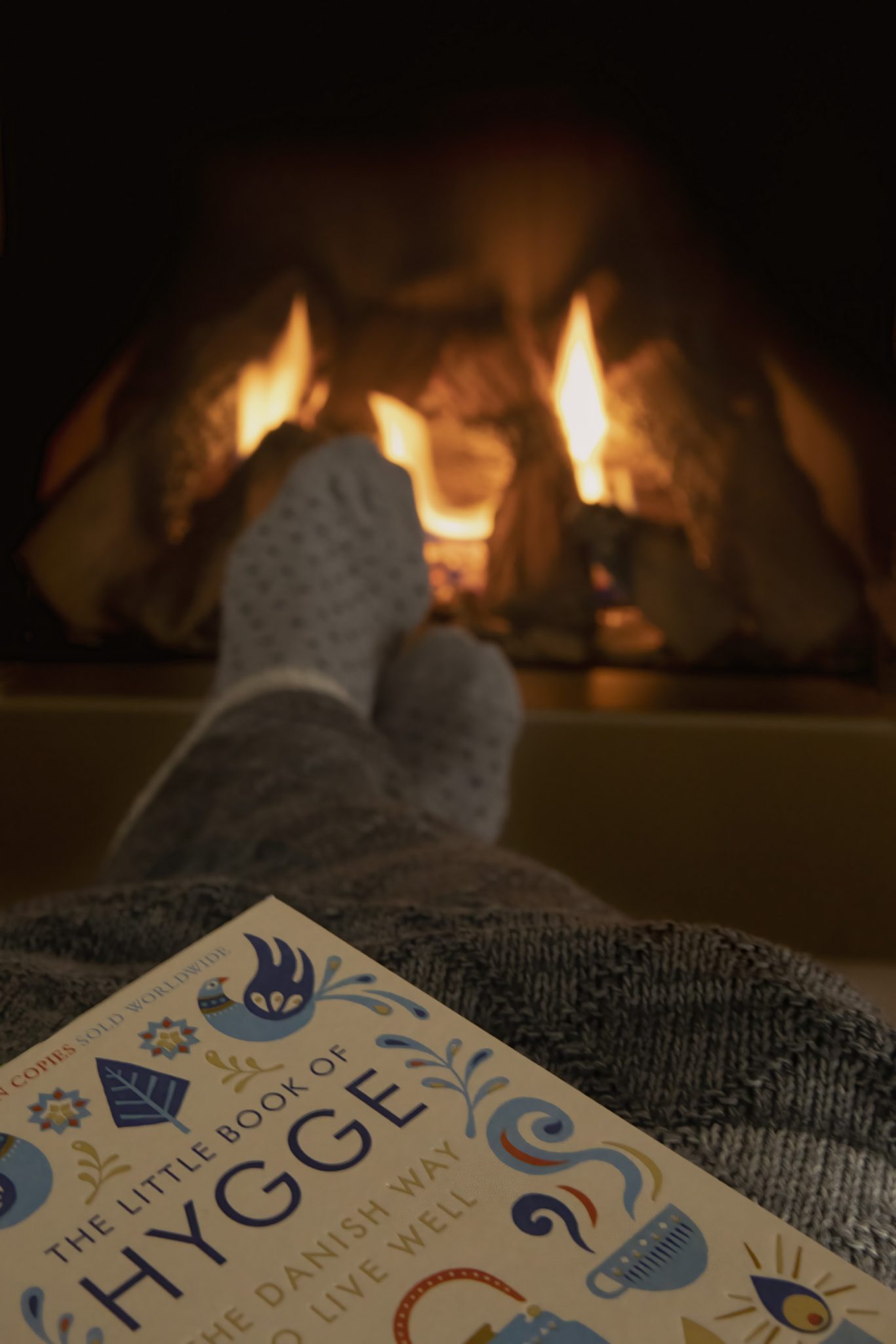 Cozy scene featuring a warm fireplace, with a pair of feet in cozy socks resting on a textured blanket, and the book titled "The Little Book of Hygge" in the foreground.