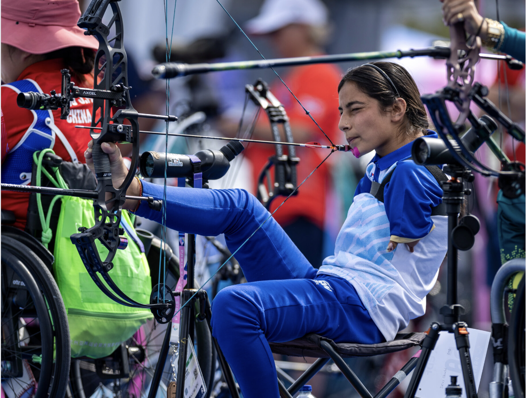 The image depicts a young girl participating in an archery competition, specifically seated archery. In the foreground, the girl is wearing a blue and white shirt with long sleeves and blue pants, sitting on a black chair designed for people with disabilities. She has dark hair tied back in a ponytail and is holding a bow with her left hand, while her right arm rests on her lap. The bow is attached to an arrow that points downwards. Behind the girl, several other archers are visible, all of whom are using specialized equipment designed for individuals with physical disabilities. In the background, a crowd of people can be seen watching the competition, although they are out of focus.