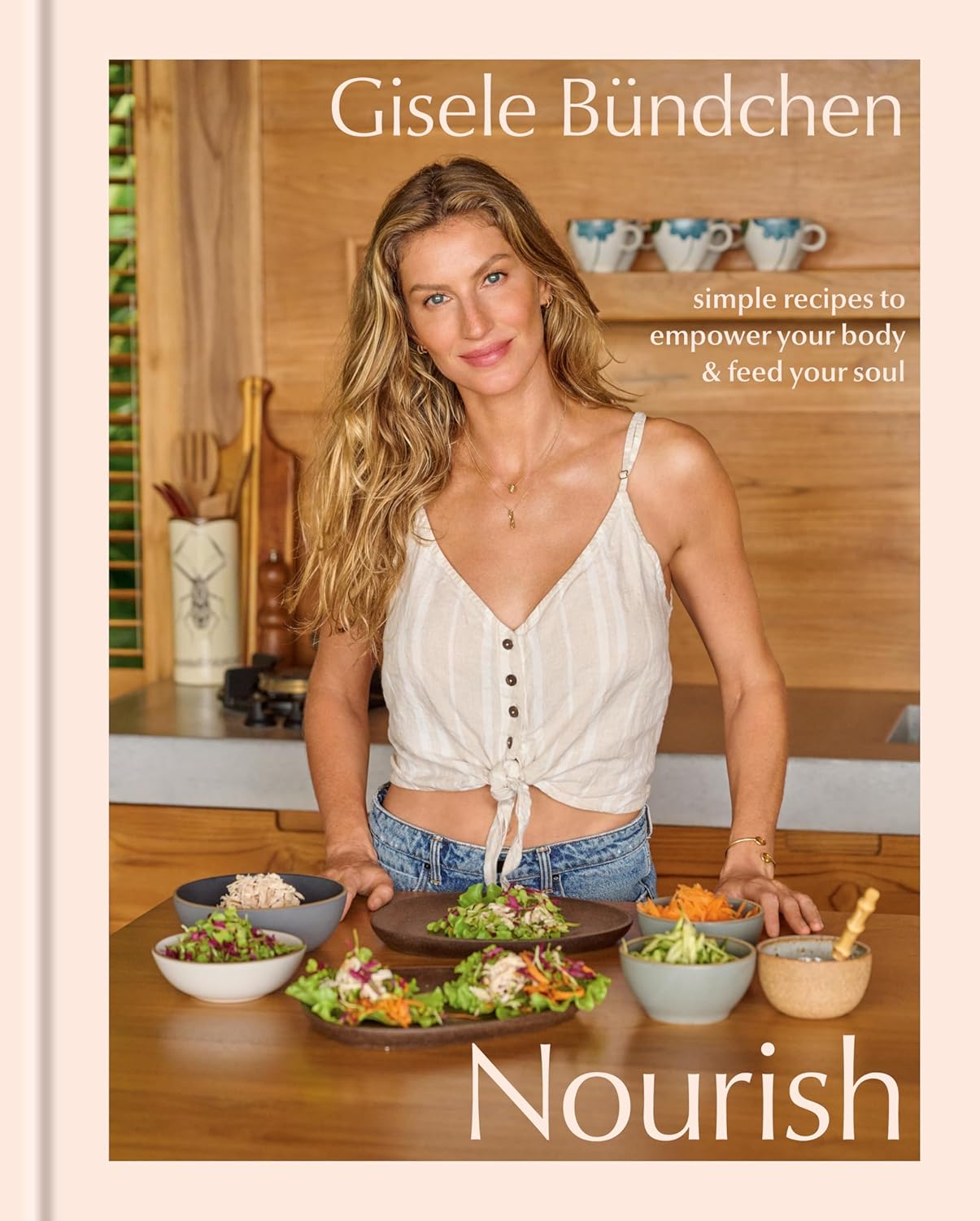 A woman with long, wavy hair smiles while standing in a kitchen, wearing a light blouse and jeans. She is surrounded by bowls of fresh salads and ingredients, with a cookbook titled "Nourish" prominently displayed. The book's subtitle reads, "simple recipes to empower your body & feed your soul."