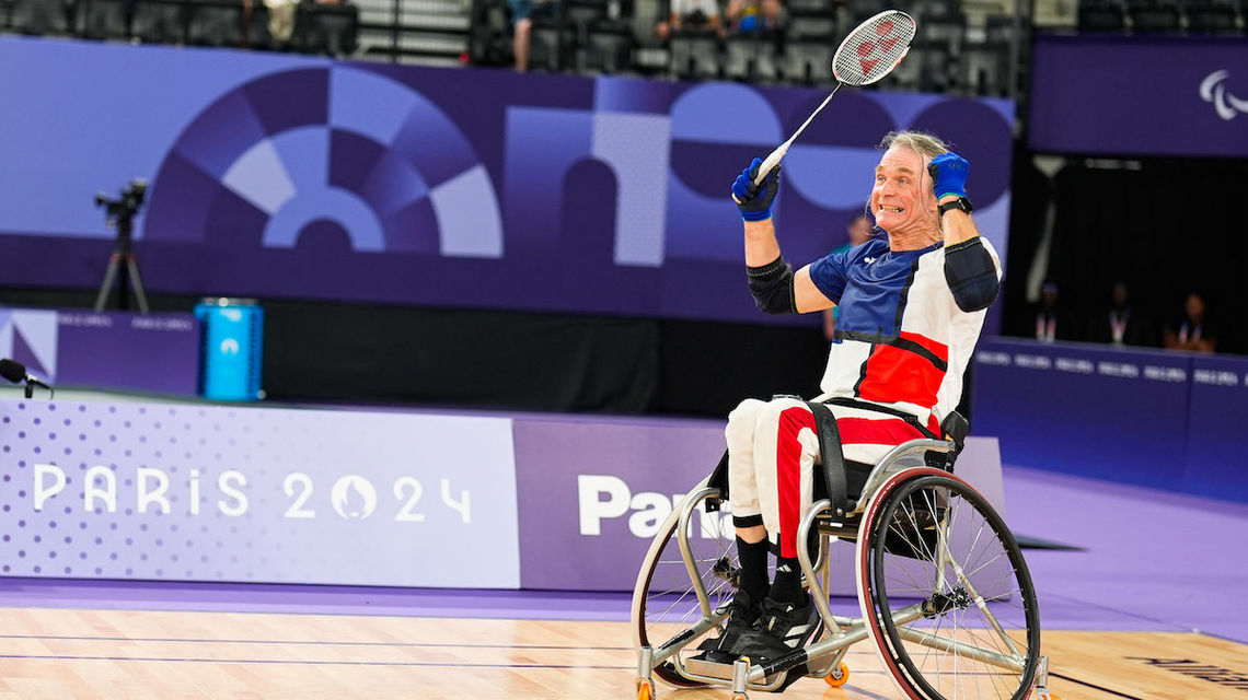 The image shows two men playing badminton in wheelchairs, with one man celebrating his victory. * The man on the left is wearing a white shirt and red shorts. * He has short gray hair and is sitting in a wheelchair. * His right arm is raised in celebration, holding a racket. * The background of this section shows a purple banner with "PARIS 2024" written in large letters. * The man on the right is also wearing a white shirt and red shorts. * He has short gray hair and is sitting in a wheelchair. * His left arm is raised in celebration, holding a racket. * The background of this section shows a purple banner with "PARIS 2024" written in large letters. The image suggests that the two men are competing in a badminton tournament for athletes with disabilities.