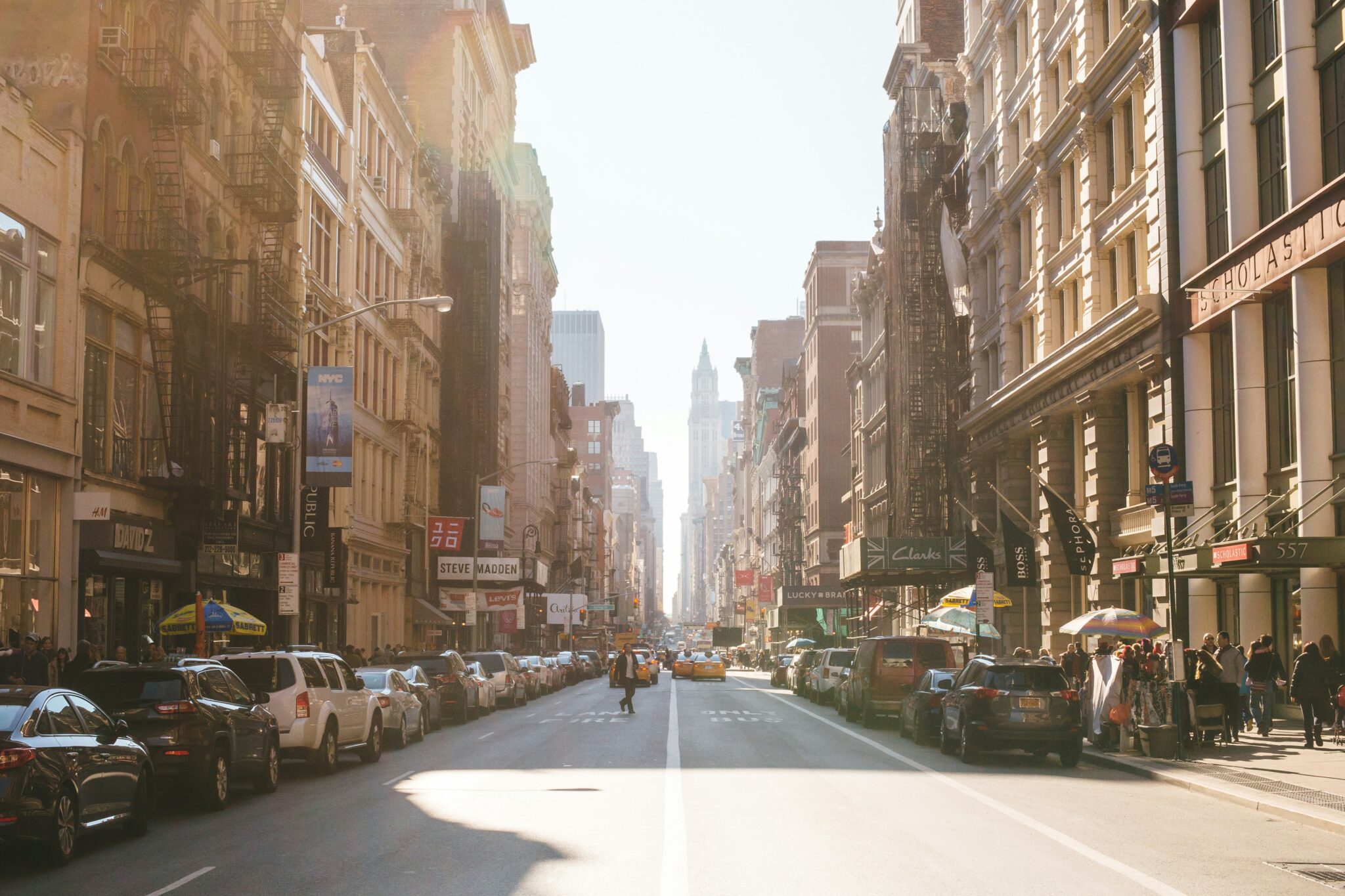 Alt tag: "Sunlit street scene in a bustling city, featuring parked cars along both sides, storefronts with signage, and a pedestrian crossing the road, framed by tall buildings in the distance."