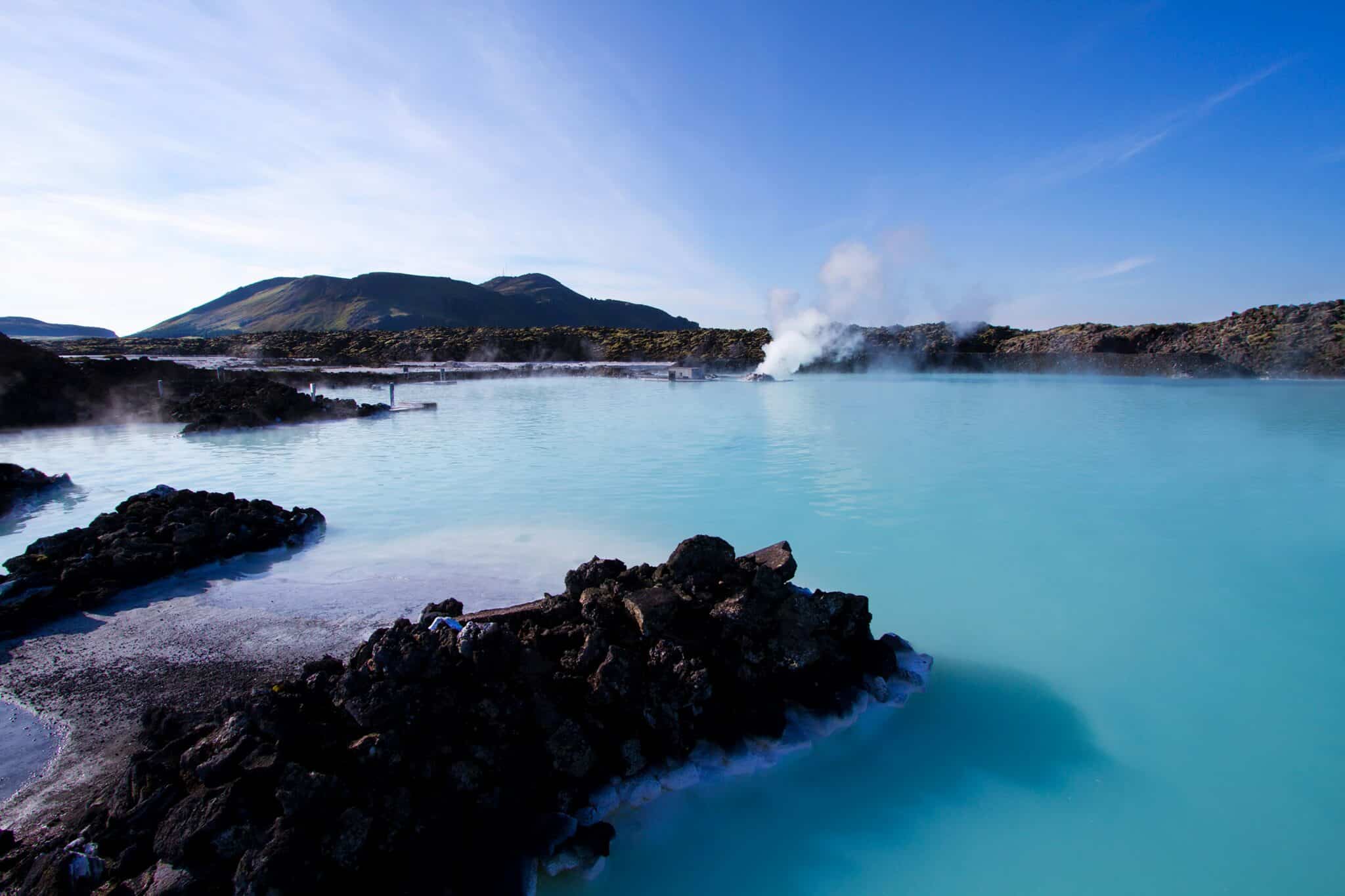 The image depicts a serene and breathtaking landscape, showcasing the iconic Blue Lagoon in Iceland. In the foreground, a large body of water dominates the scene, with its vibrant blue hue reflecting the surrounding environment. The water's surface is dotted with small waves, adding to the sense of movement and life. A cluster of rocks protrudes from the water, providing a natural barrier between the lagoon and the adjacent landmass. The background features a majestic mountain range, characterized by gentle slopes and lush greenery. The sky above is a brilliant blue, with wispy clouds scattered throughout, creating a sense of depth and atmosphere. The overall ambiance of the image exudes tranquility and beauty, inviting the viewer to immerse themselves in the natural wonder of the Blue Lagoon.