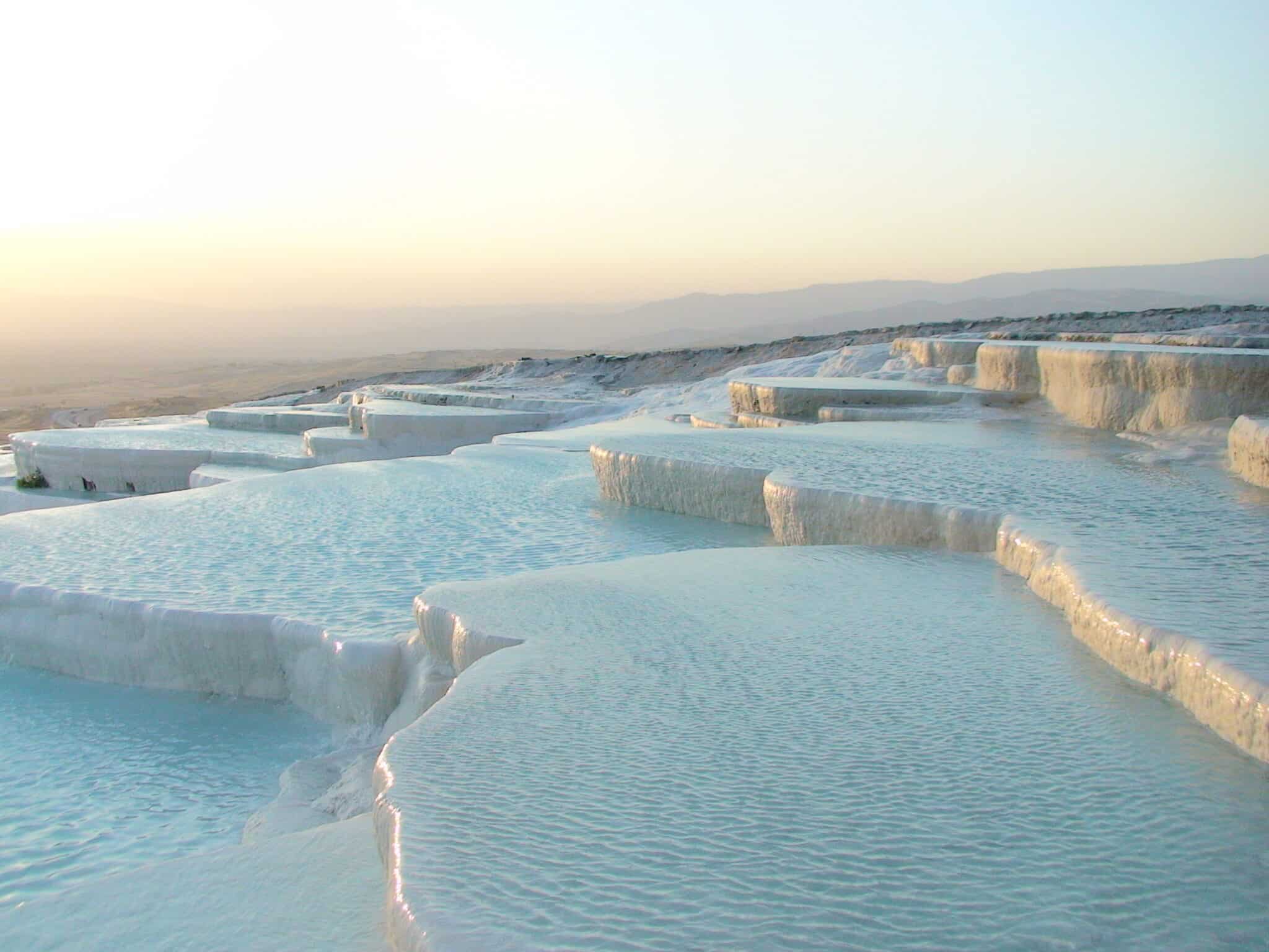This photograph showcases a unique landscape featuring white, stepped terraces of travertine formations in the foreground and background. The foreground features numerous rectangular-shaped pools filled with clear blue water, while the background showcases several parallel rows of flat, white stone-like structures that resemble steps or platforms. The terrain slopes gently upward toward the horizon, where additional rows are visible on either side. In the distance, a mountain range stretches across the top-right corner of the image, set against an overcast sky with a yellow hue at the horizon and a blue tone above.