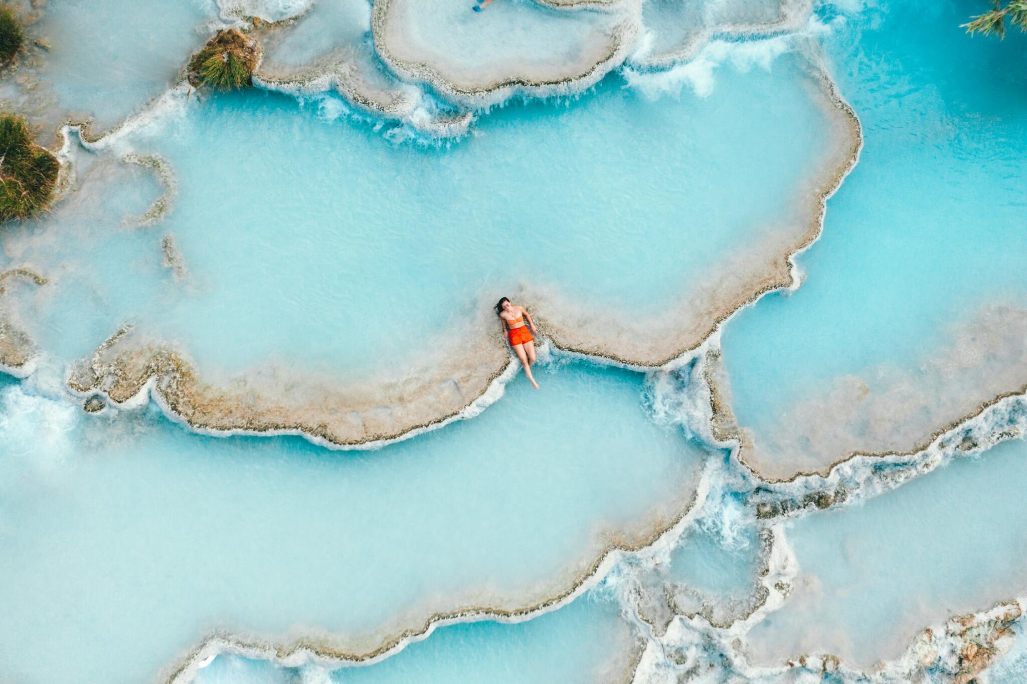 Aerial view of a person in a bright orange swimsuit relaxing in azure thermal pools with unique, layered formations.