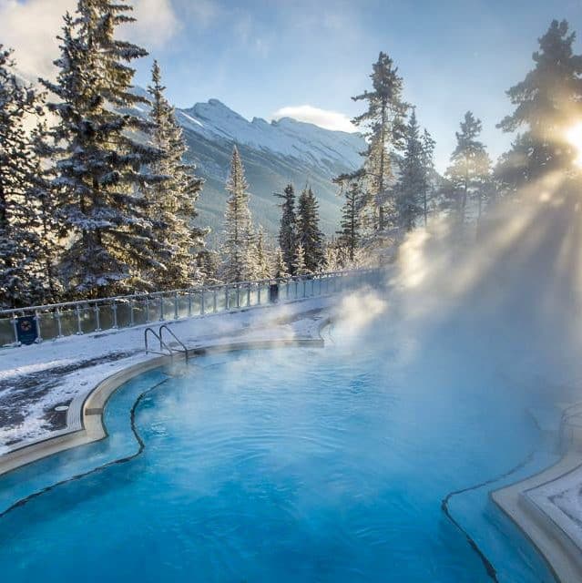 The image presents a serene winter scene, featuring a large outdoor pool situated in the snow-covered mountains. In the foreground, the pool is surrounded by evergreen trees and appears to be heated as steam rises from its surface. The surrounding landscape is blanketed with snow, while the sun shines brightly above the mountain range in the background. A fence runs along the edge of the pool, separating it from the surrounding terrain. The overall atmosphere of the image exudes tranquility and relaxation, suggesting a perfect setting for a winter getaway or vacation.