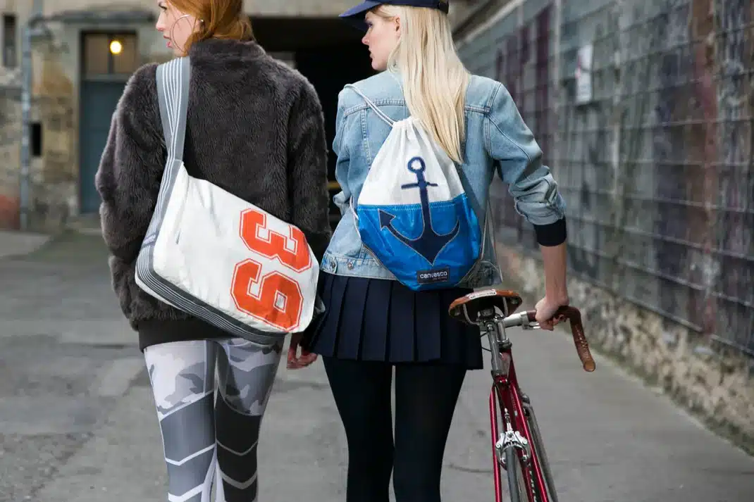 The image shows two women walking down a street, with one of them carrying a drawstring bag and the other holding a bicycle. * Two women: + One woman has blonde hair and is wearing a blue baseball cap, a denim jacket, black leggings, and a white and blue drawstring bag with an anchor design on it. + The other woman has red hair and is wearing a gray fur coat, a white and orange number 33 jersey, and camouflage pants. * A bicycle: + It is being held by the blonde-haired woman in her right hand. + The bike has a red frame and silver handlebars. * A drawstring bag: + It is being carried by the blonde-haired woman over her left shoulder. + The bag is white with a blue anchor design on it. The image appears to be an advertisement for the drawstring bag, showcasing its unique design and versatility.