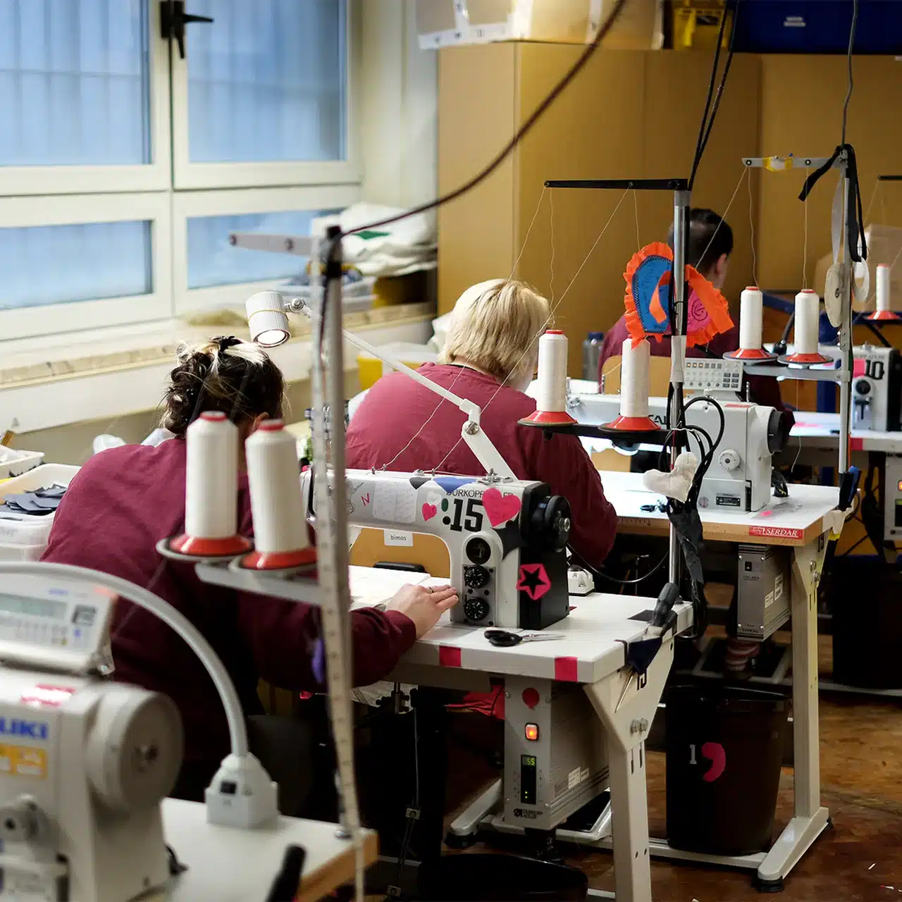 The image depicts a group of individuals engaged in sewing or tailoring activities within a workshop setting. In the foreground, several sewing machines are prominently displayed on tables, each accompanied by spools of thread and other necessary equipment. The individuals are dressed in red long-sleeved shirts with white logos on their backs, suggesting they may be employees or apprentices working together in a team environment. The background features two windows along one wall, allowing natural light to illuminate the space and providing a glimpse into the outside world. The overall atmosphere appears to be one of focused work and collaboration, with the individuals intently concentrating on their tasks as they contribute to the creation of various garments or textiles.